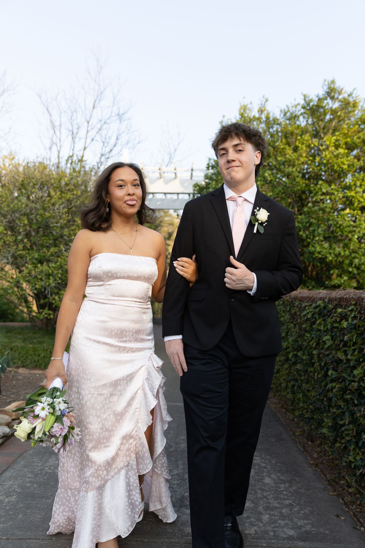 A young couple dressed in formal attire walk together outdoors, she in a white gown, he in a black suit with boutonniere.