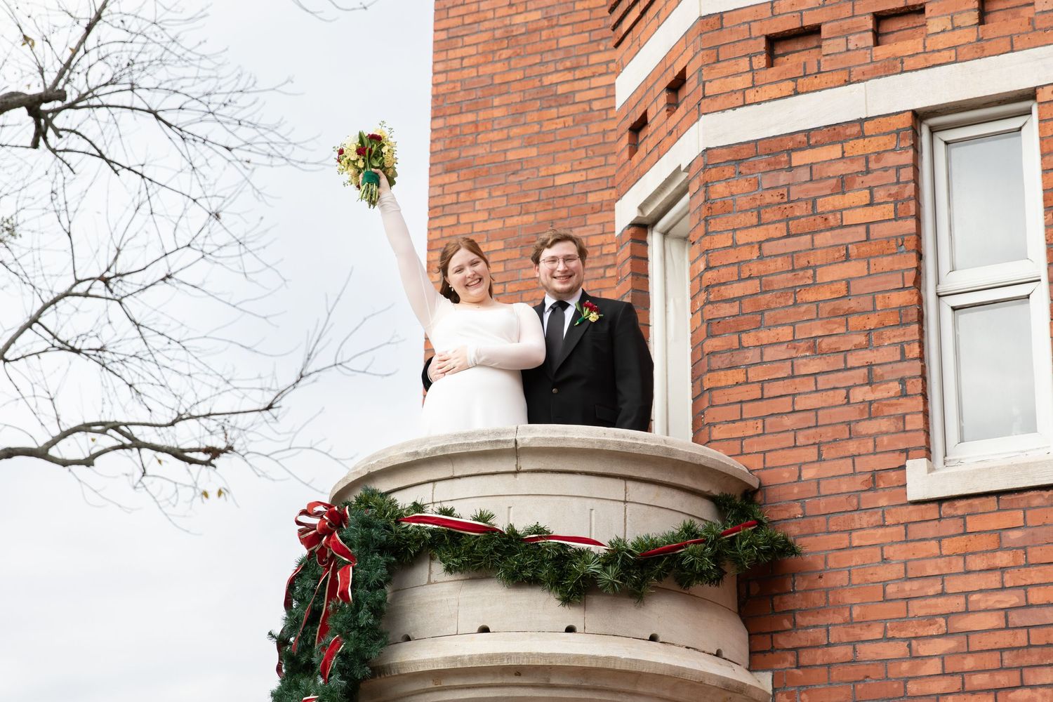 Bride and groom celebrate on a brick building balcony decorated with Christmas garland, bride raising bouquet.