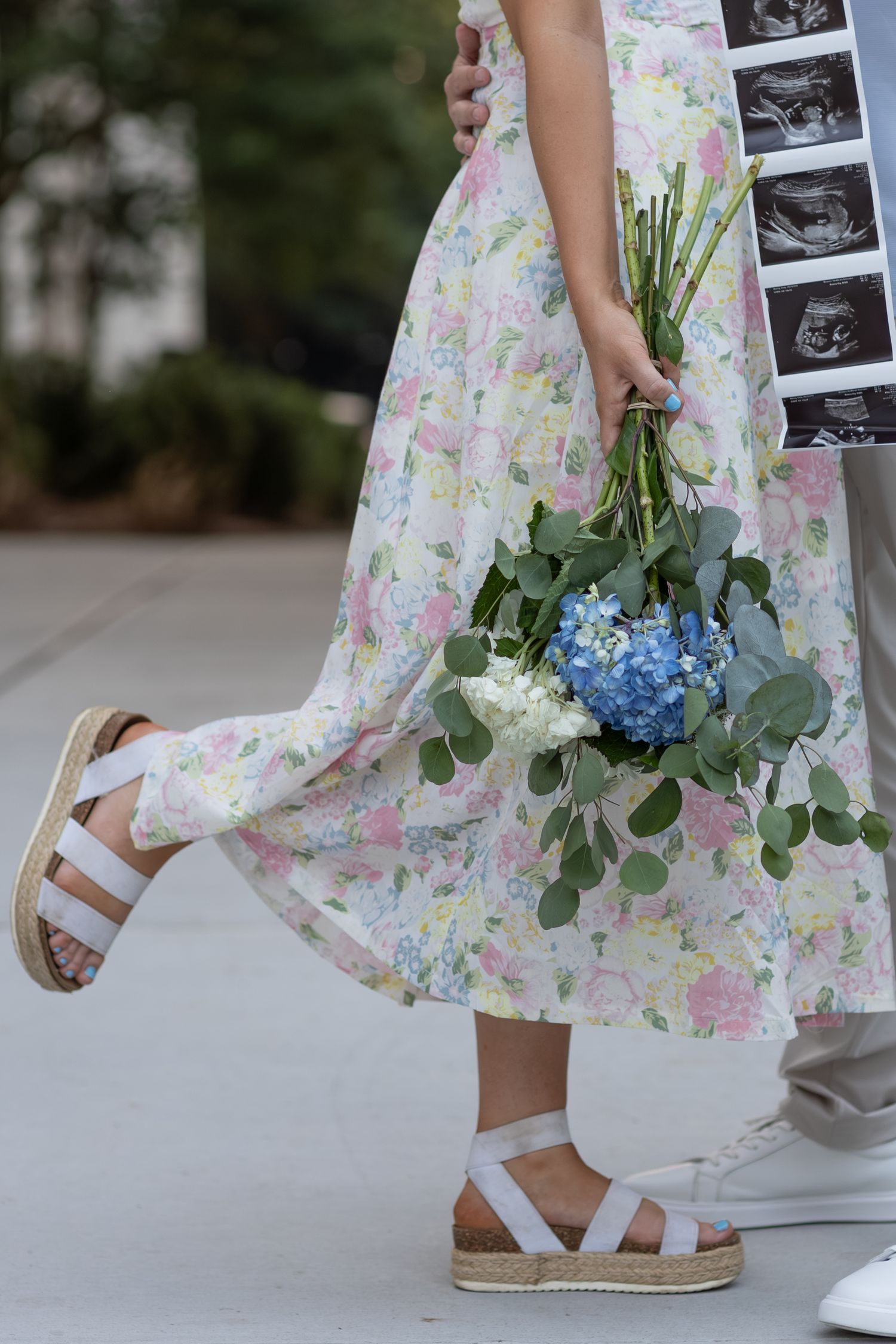 Woman in floral dress holding blue hydrangea bouquet with eucalyptus, wearing white platform sandals, carrying ultrasound photos.