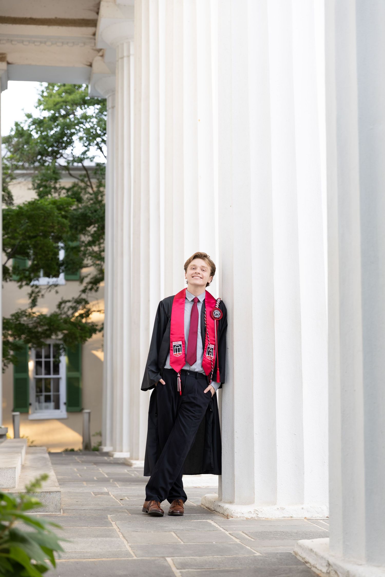 Graduate in black gown and red sash leans against white columns at a university building.