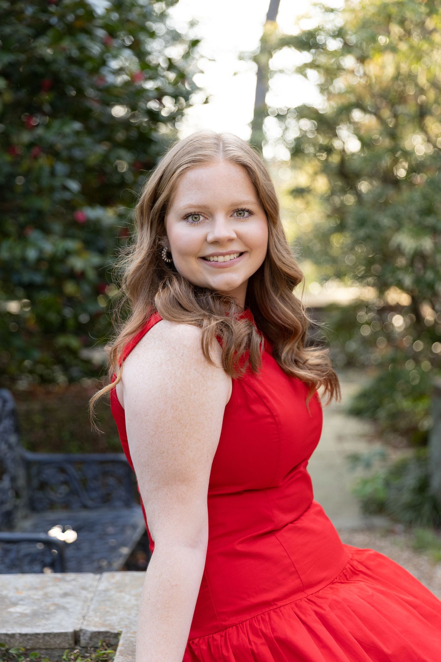 Young woman in red dress smiling outdoors in garden setting with green foliage background.