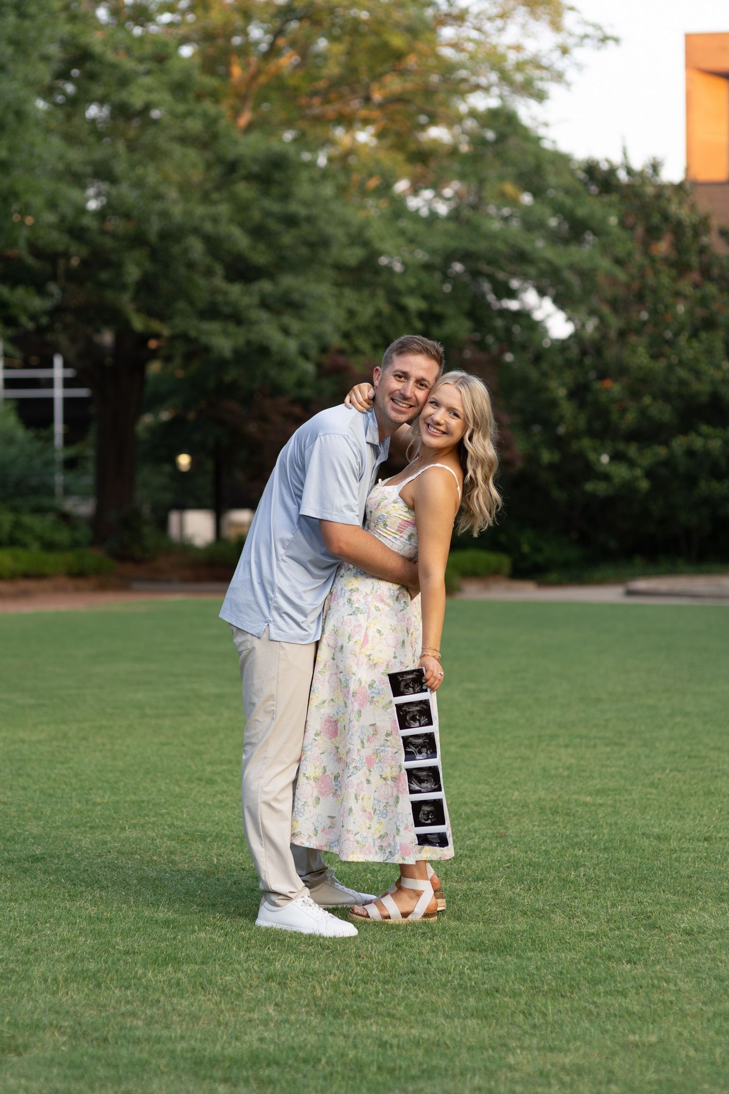Smiling couple stands on green lawn holding ultrasound photos for pregnancy announcement.