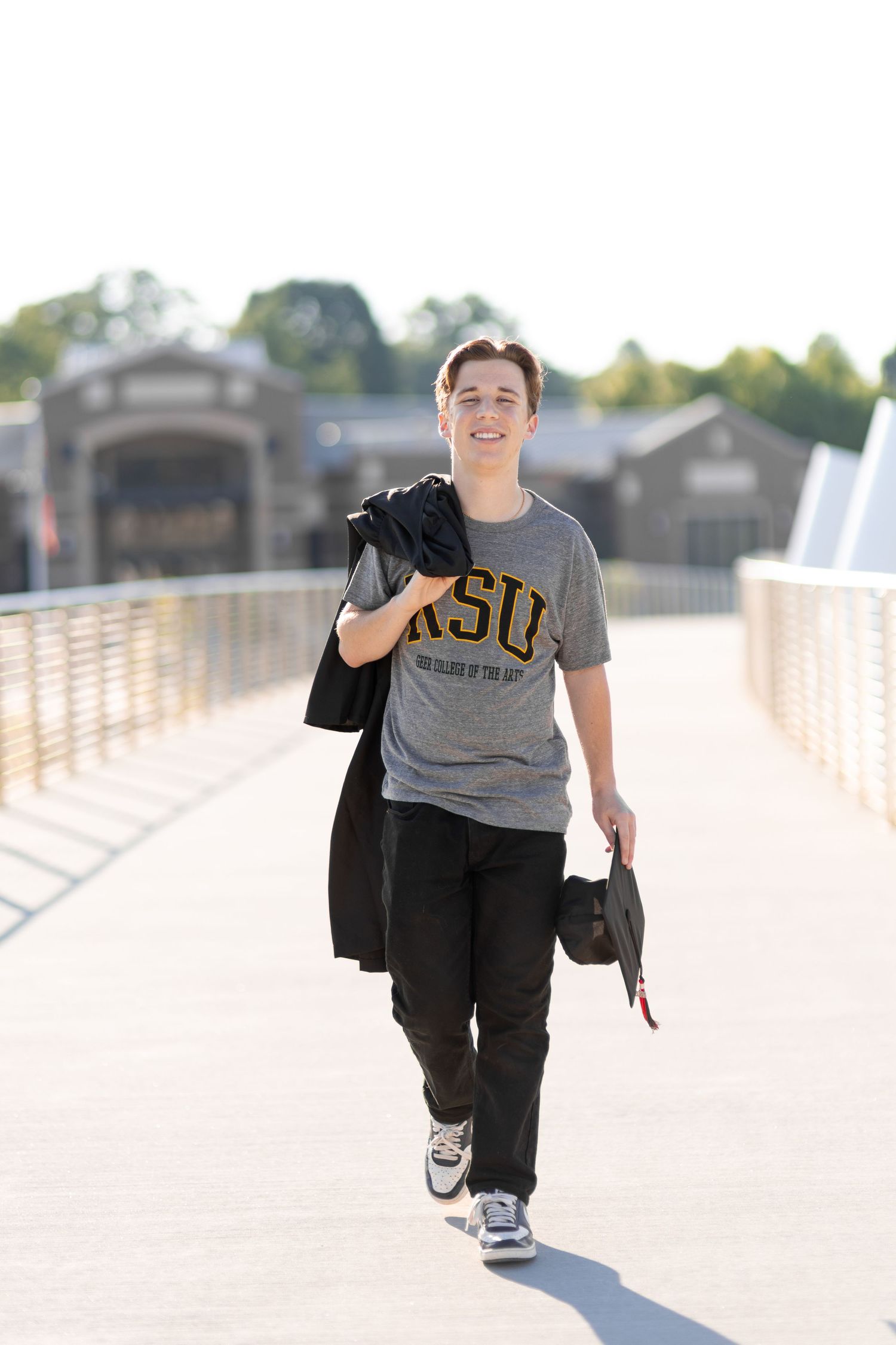 Smiling young man walking on a bridge wearing a gray KSU t-shirt, carrying a black jacket and graduation cap.