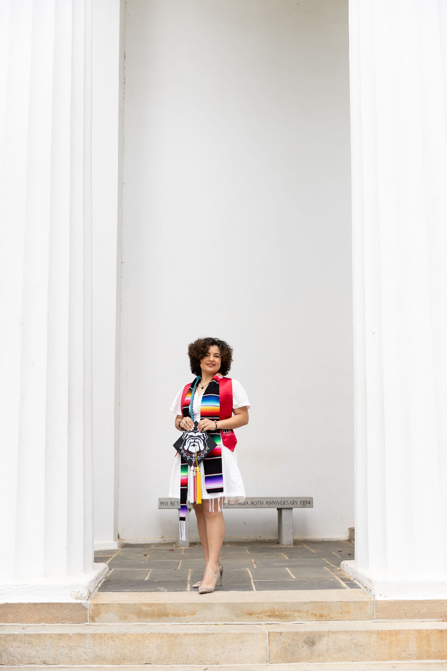 Woman in colorful striped dress holding patterned bag stands between white columns on stone steps.