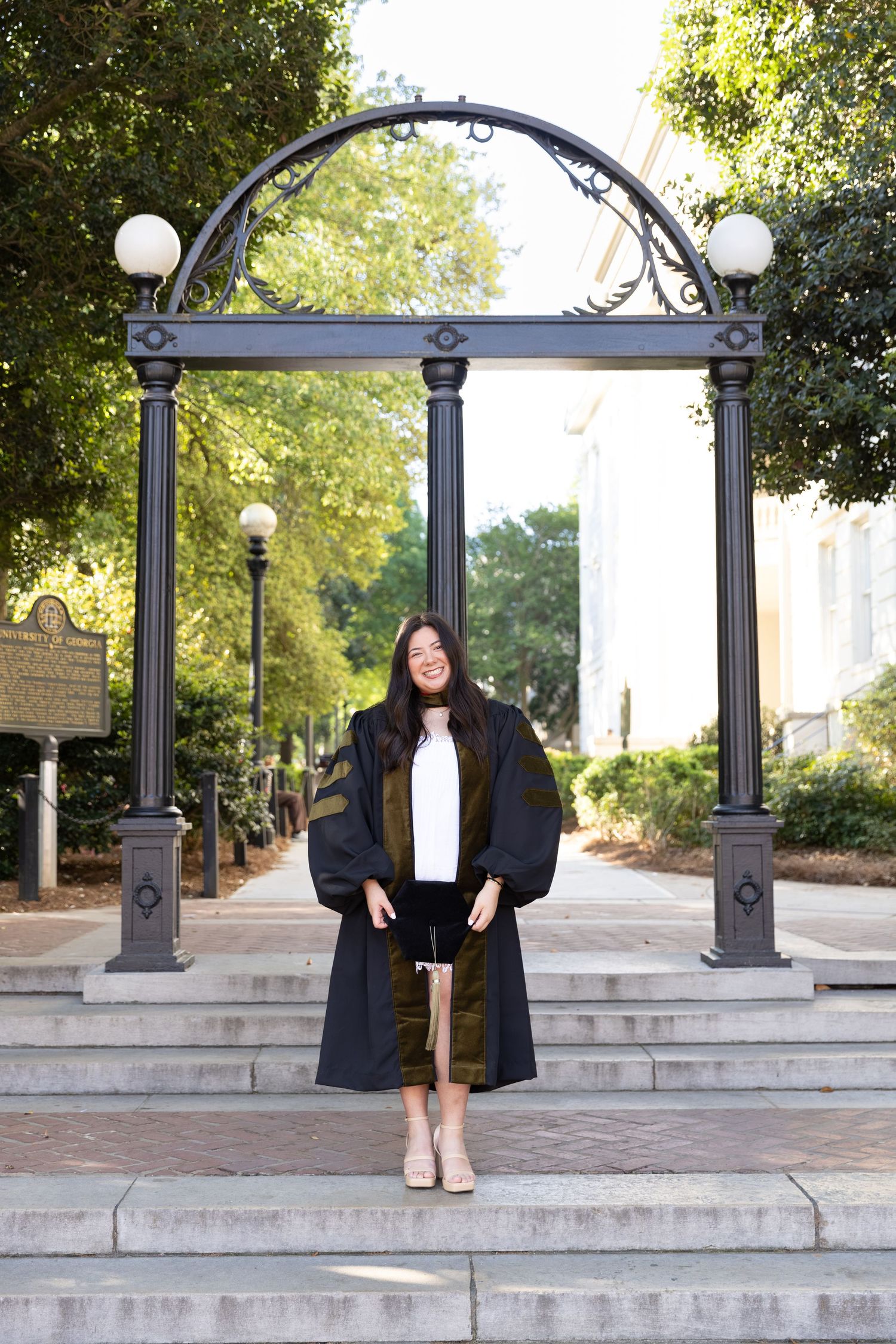 Smiling graduate in black cap and gown standing before iconic arch on university campus steps.