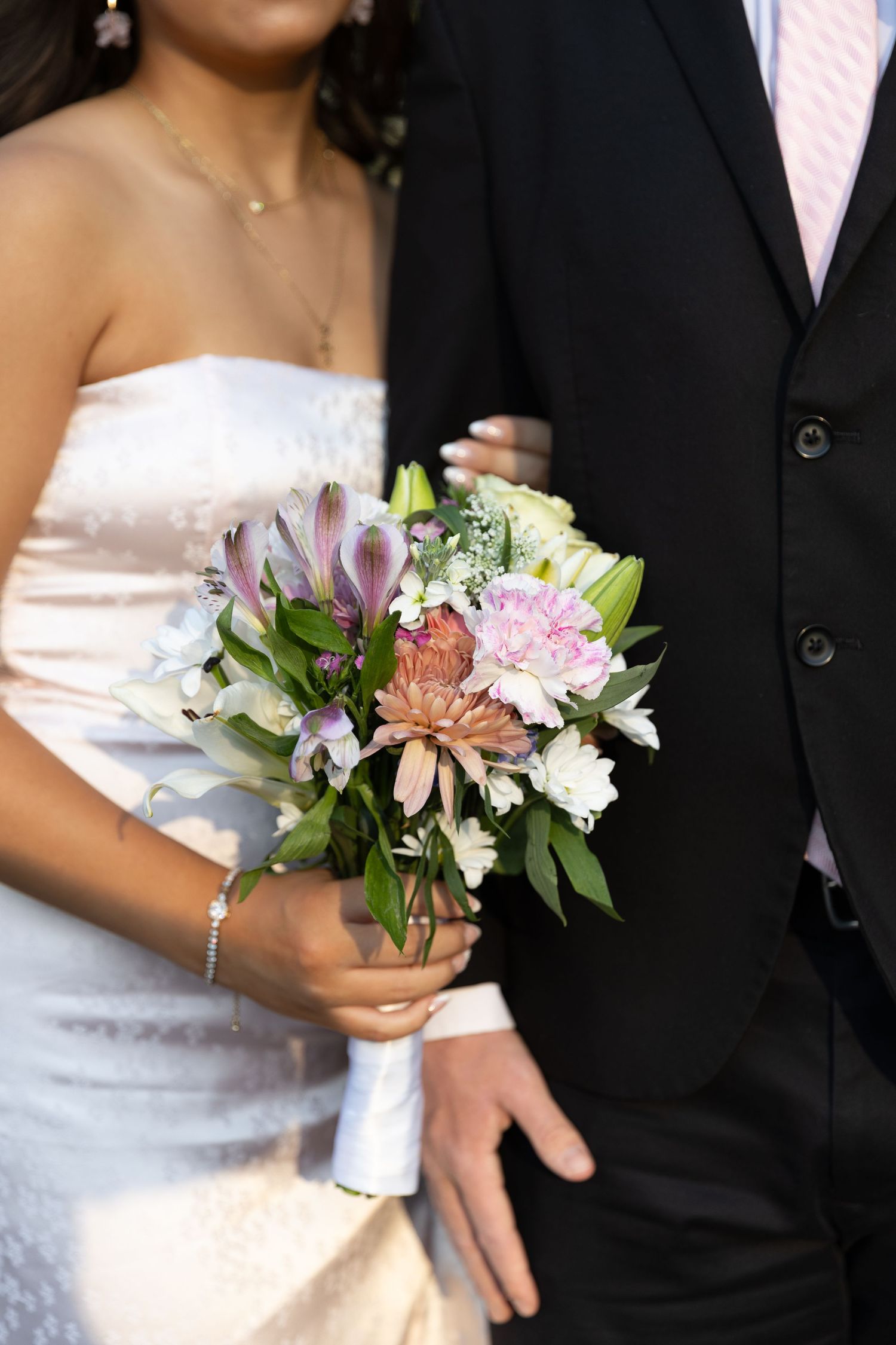 Bride in white strapless gown holds colorful bouquet with pink and purple flowers beside groom in black suit.
