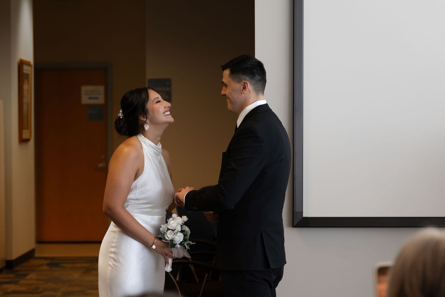 Bride in white dress laughing with groom in black suit during intimate wedding ceremony indoors.