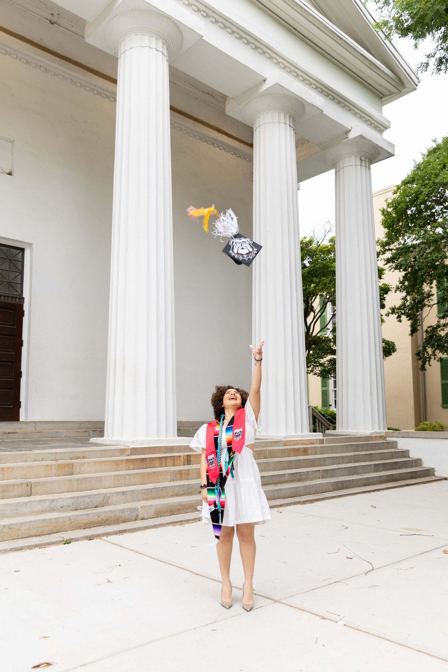 Graduate in white dress tosses cap before neoclassical building with white columns, wearing colorful stoles.