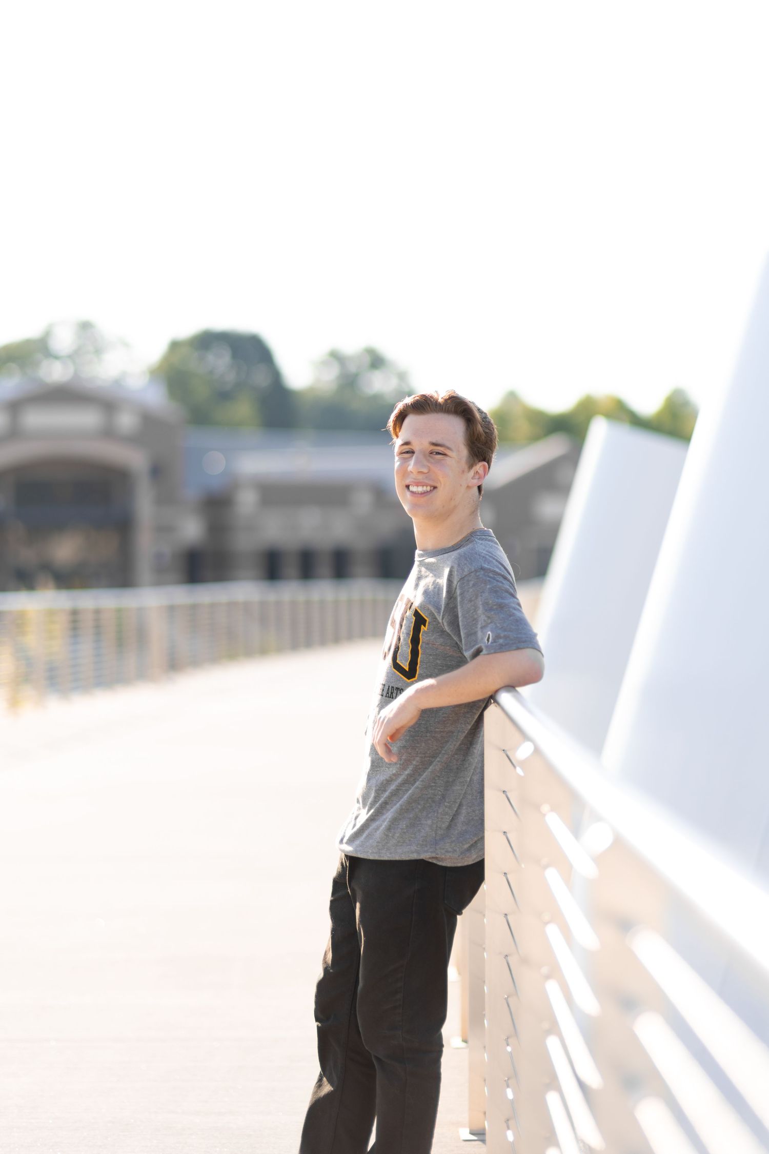 A smiling teenage boy in a gray t-shirt leans against a white bridge railing outdoors on a sunny day.