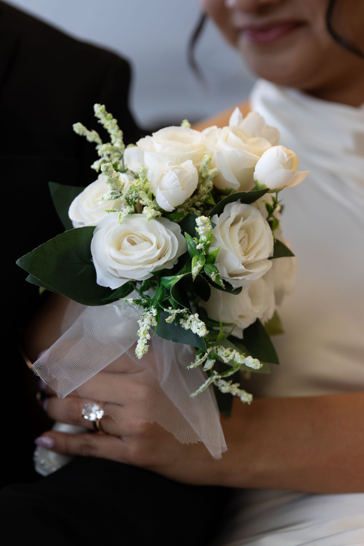 Bride holding a white rose bouquet with tulle ribbon, wearing a diamond ring, in a white wedding dress.