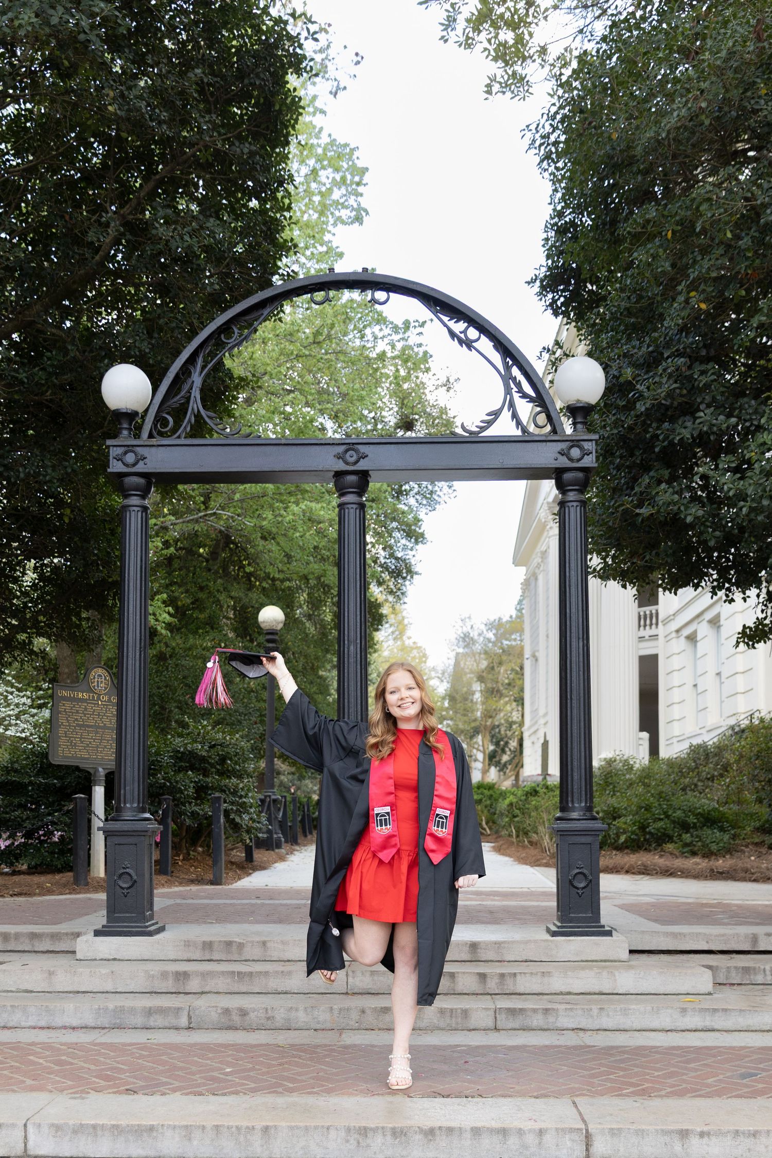 A smiling graduate in black gown and red dress poses under the UGA Arch holding a red tassel on brick steps.