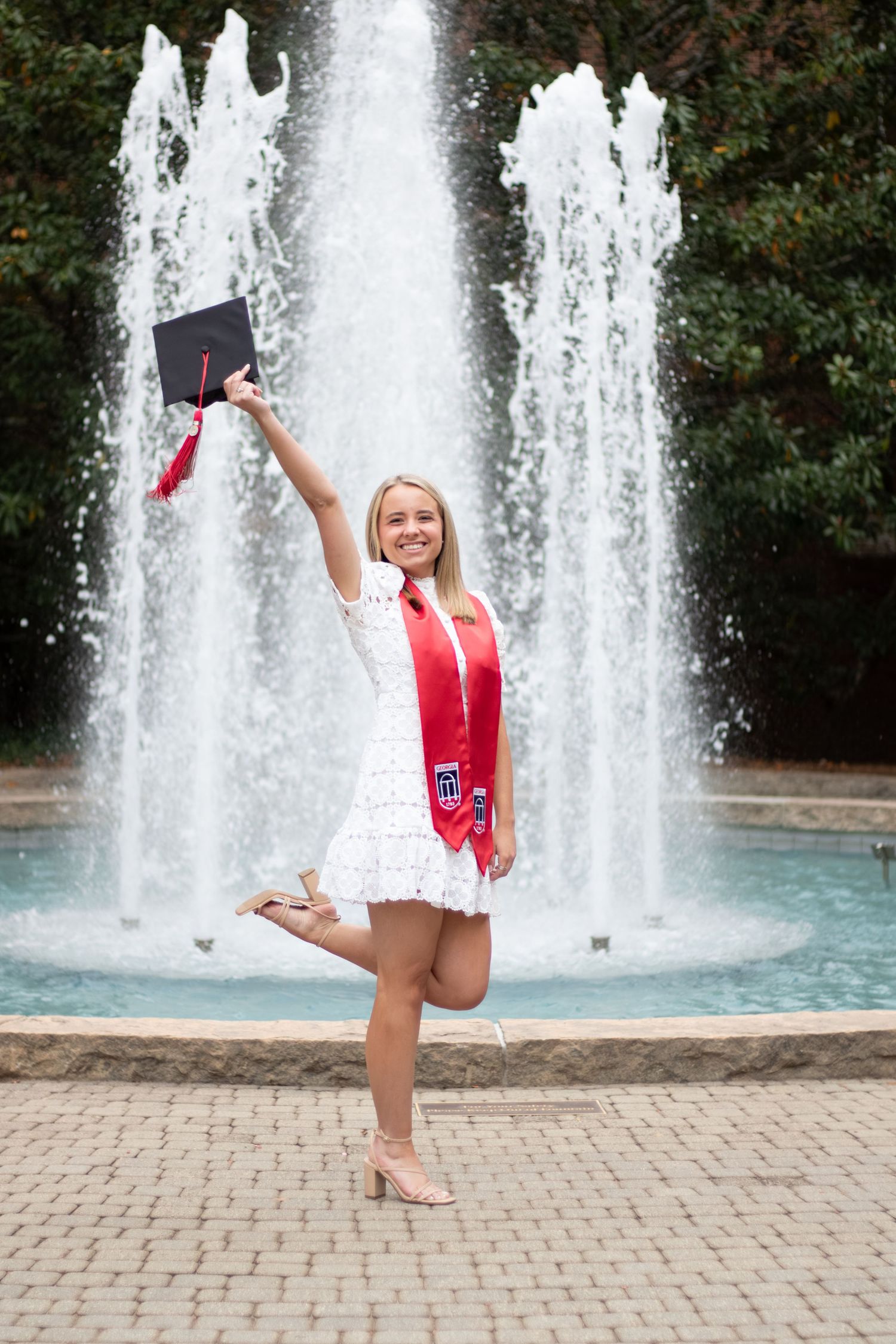 Excited college graduate in white dress and red stole holds cap up by a fountain outdoors.