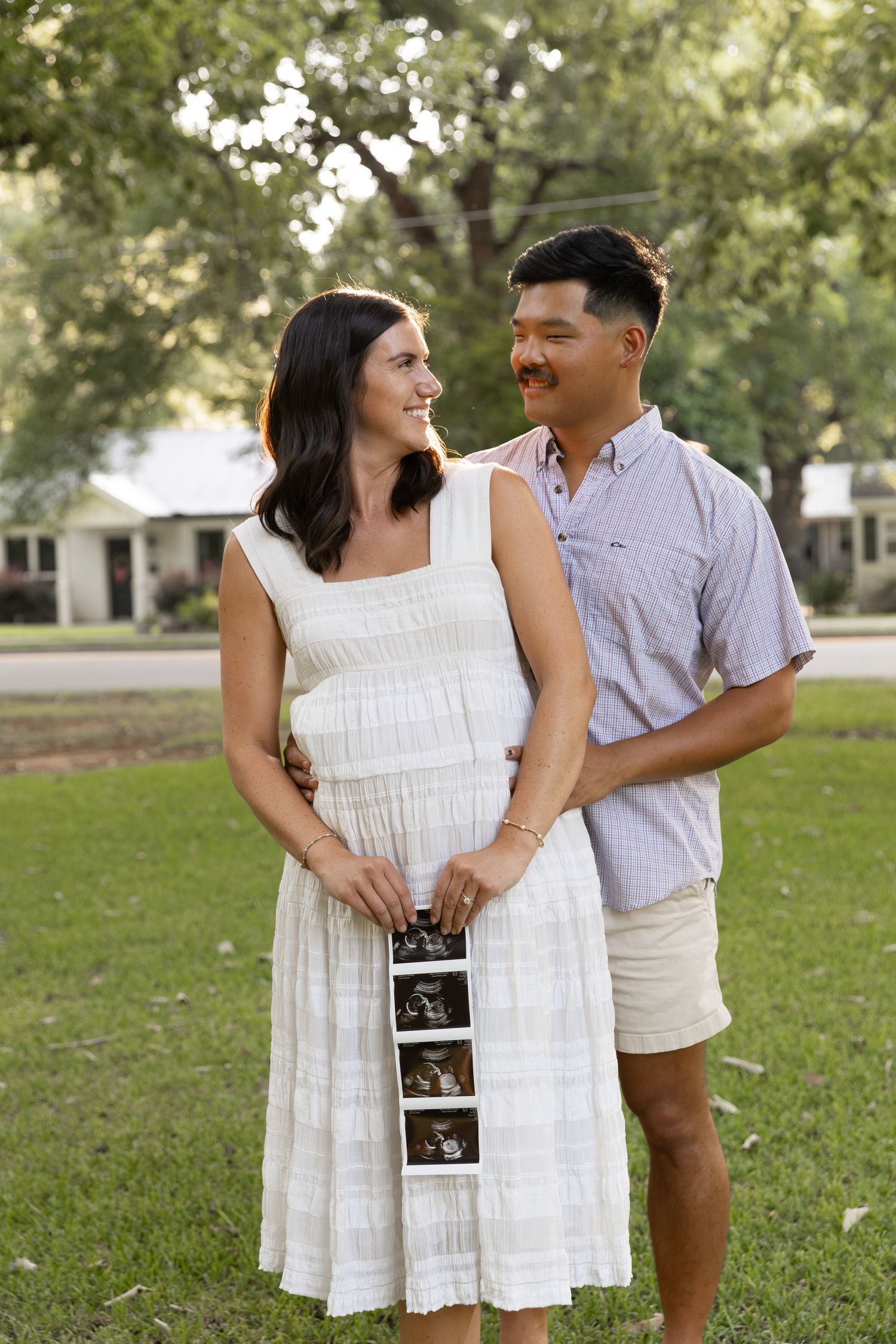 Pregnant woman in white dress holds ultrasound photos with partner outdoors in a sunny park setting.