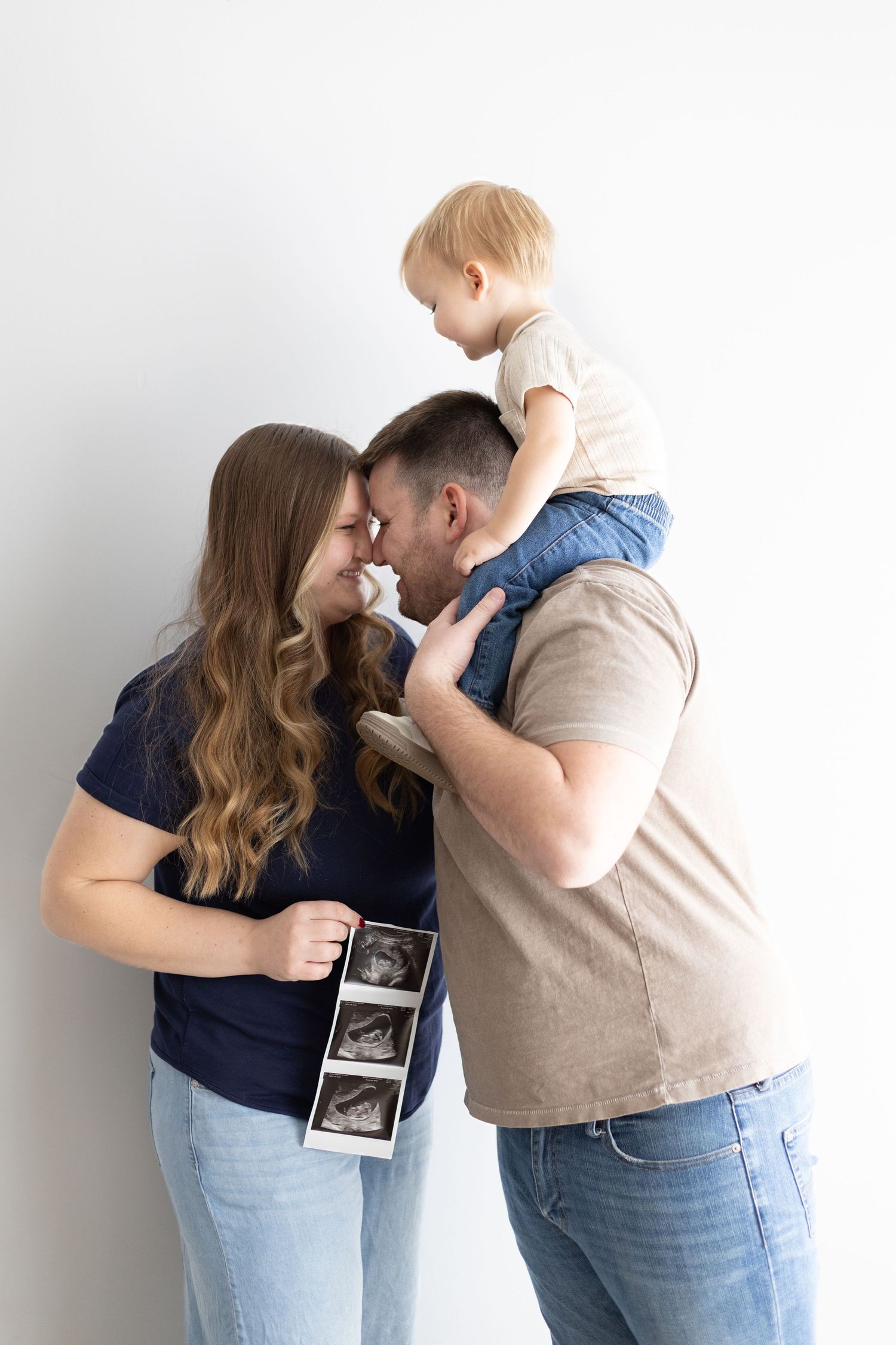 Happy couple touching noses while holding ultrasound photos, with toddler sitting on dad's shoulders against white background.
