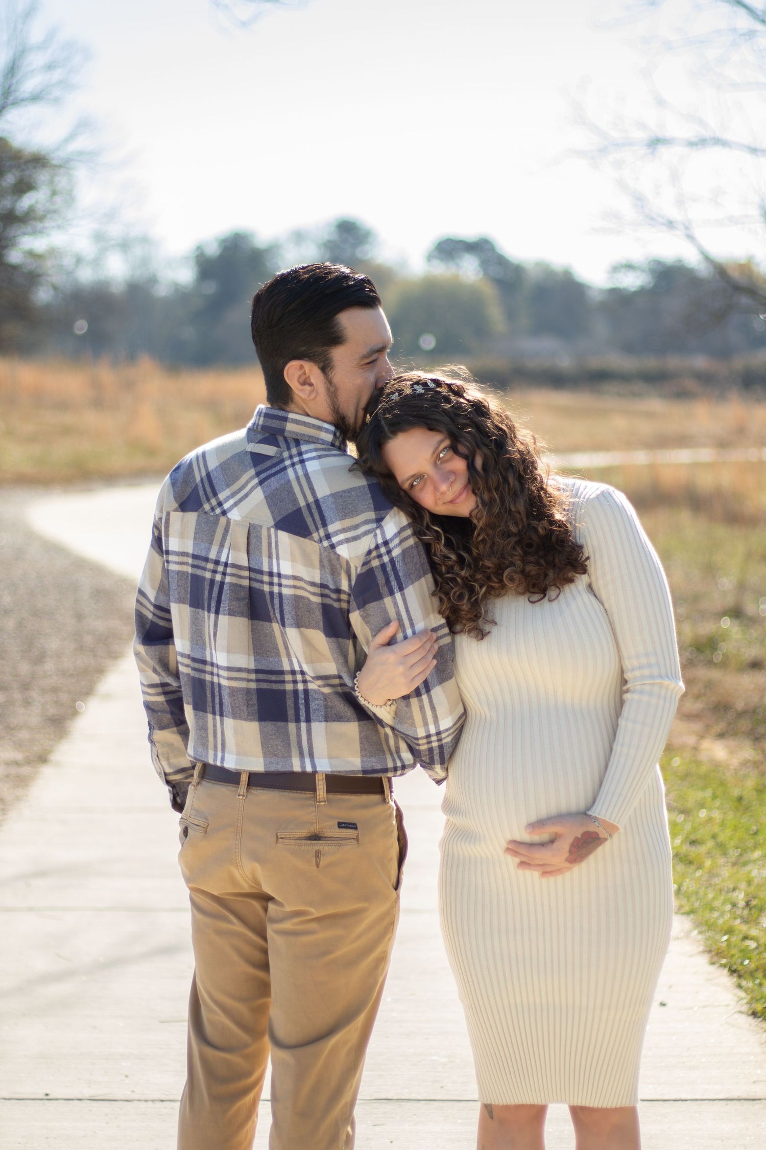 A couple shares a tender moment outdoors, the man kissing a smiling pregnant woman in a white dress on a sunny path.
