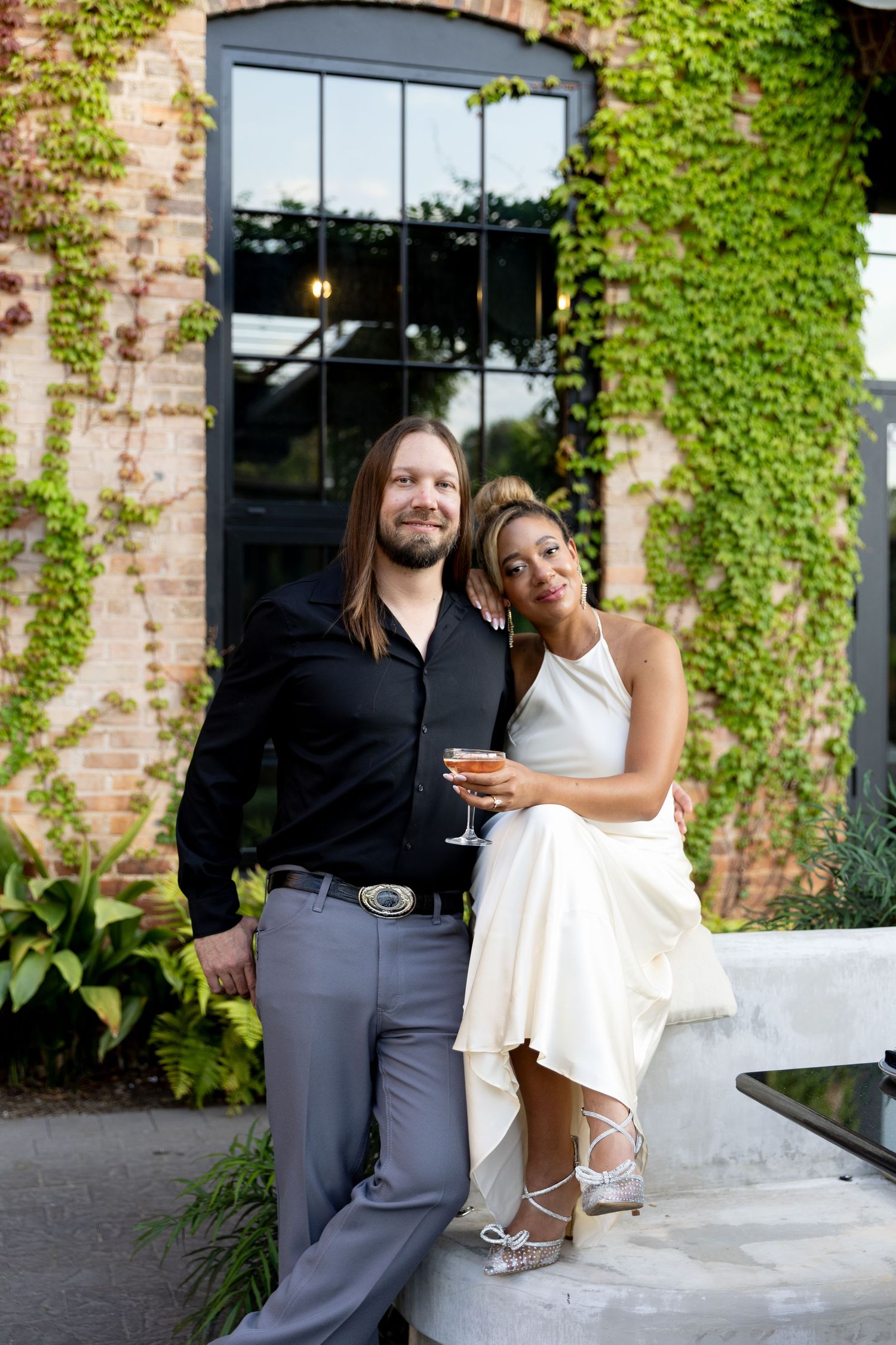 Elegant couple at outdoor venue with ivy-covered walls, woman in white dress holding champagne glass.