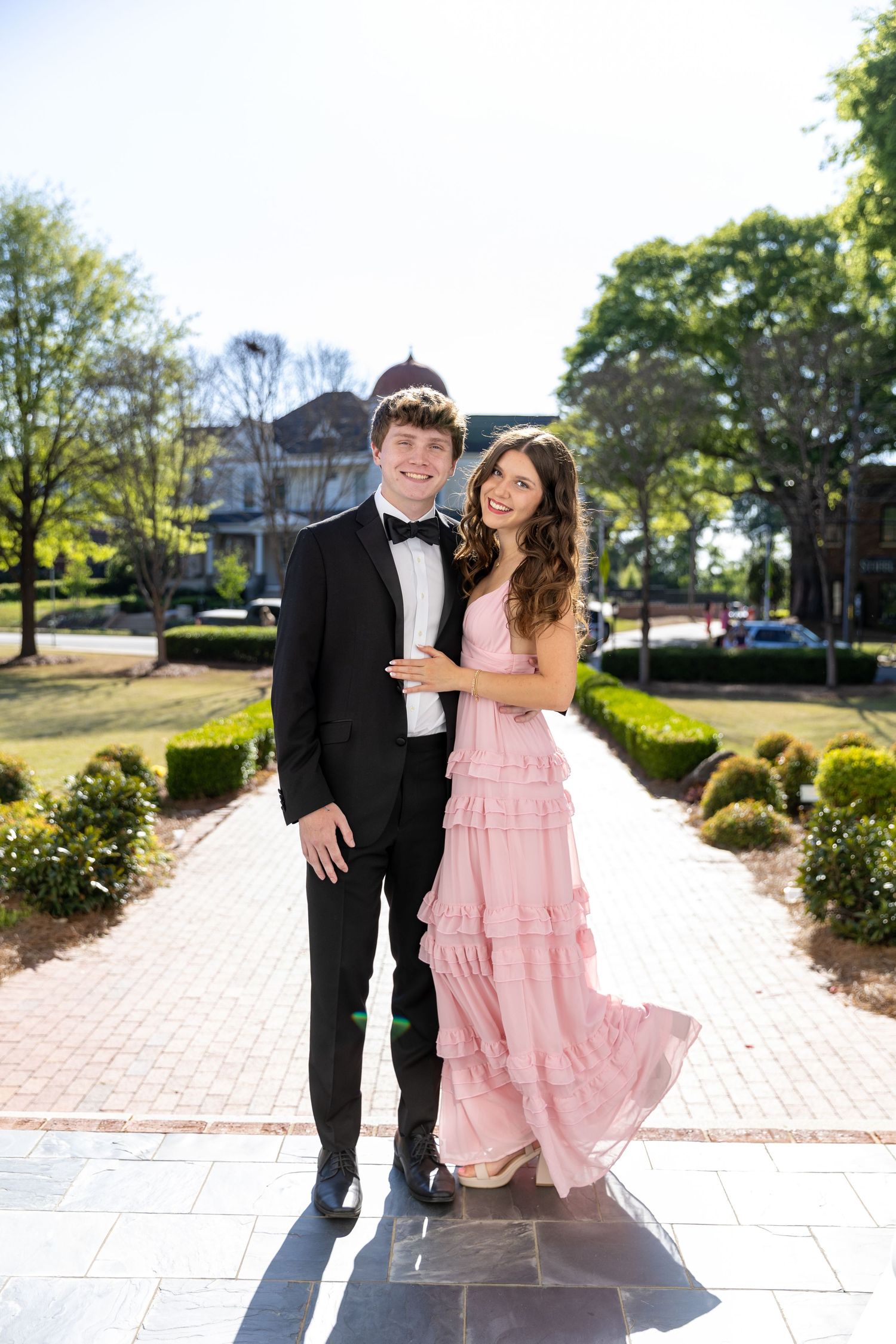 A young couple dressed in prom attire, tuxedo and pink gown, posing on a brick pathway outdoors.