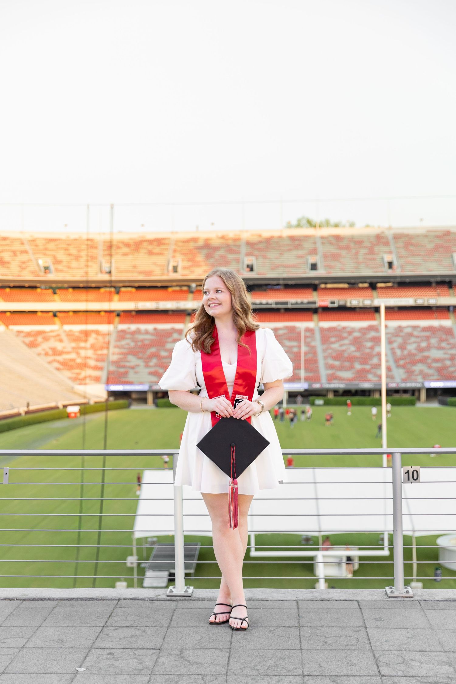 Young woman in white graduation gown with red sash holds cap at stadium, smiling proudly on graduation day.