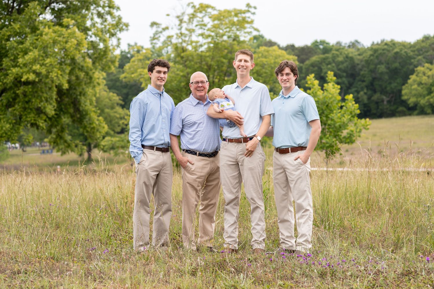 Four men posing outdoors in a grassy field, wearing matching khaki pants and blue shirts for a family portrait.