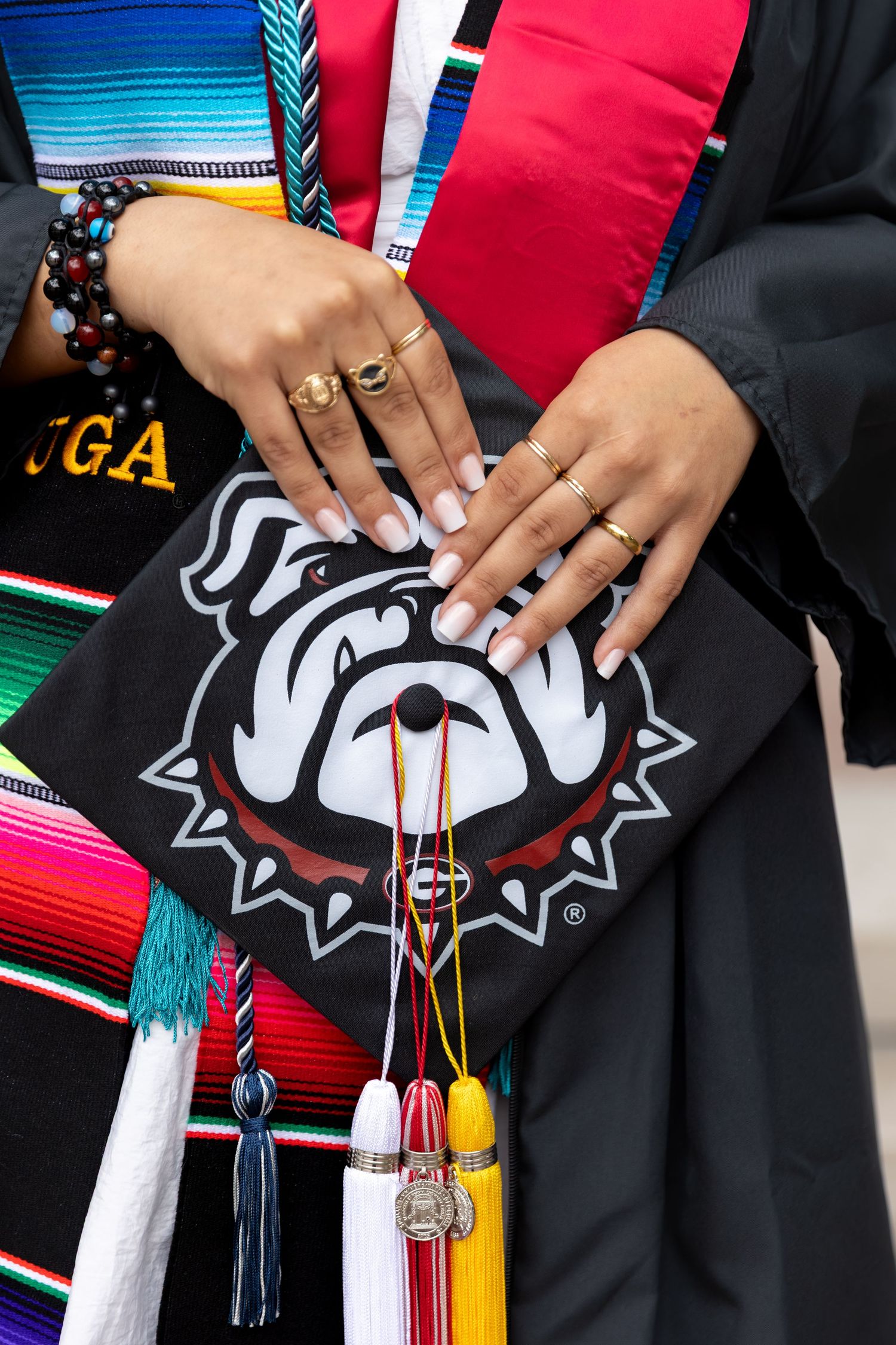 Hands with rings rest on a UGA Bulldogs graduation cap with colorful tassels and stoles.