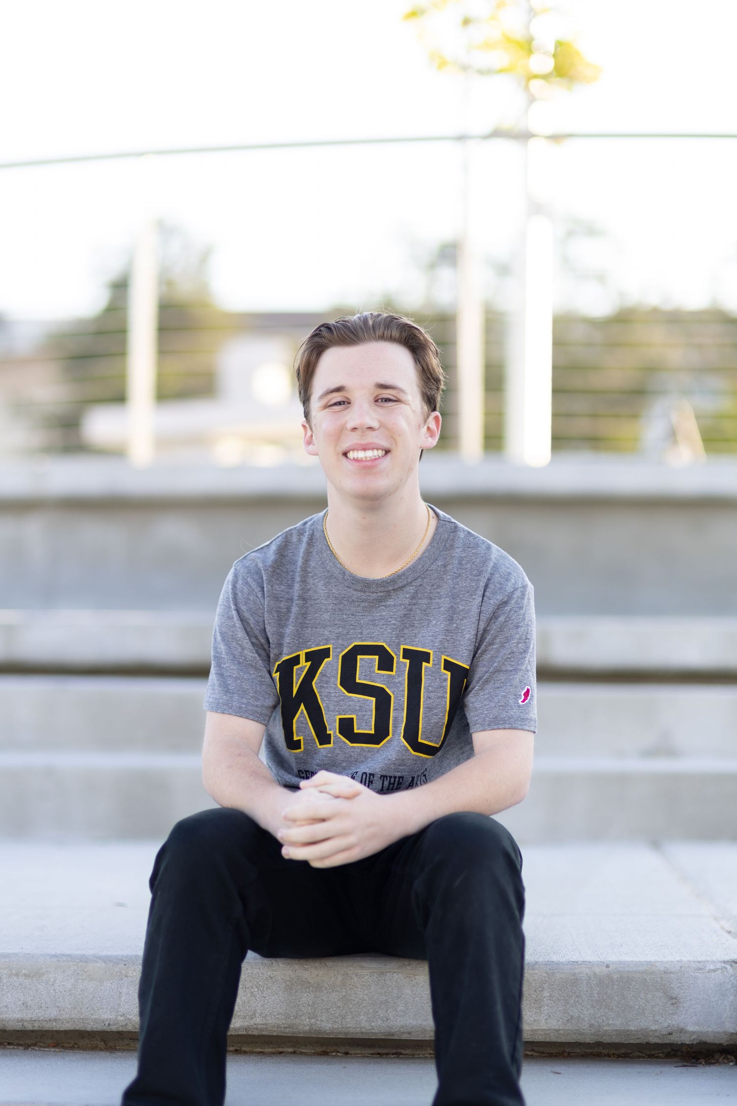 Smiling young man in KSU gray t-shirt sitting on outdoor steps with hands clasped.