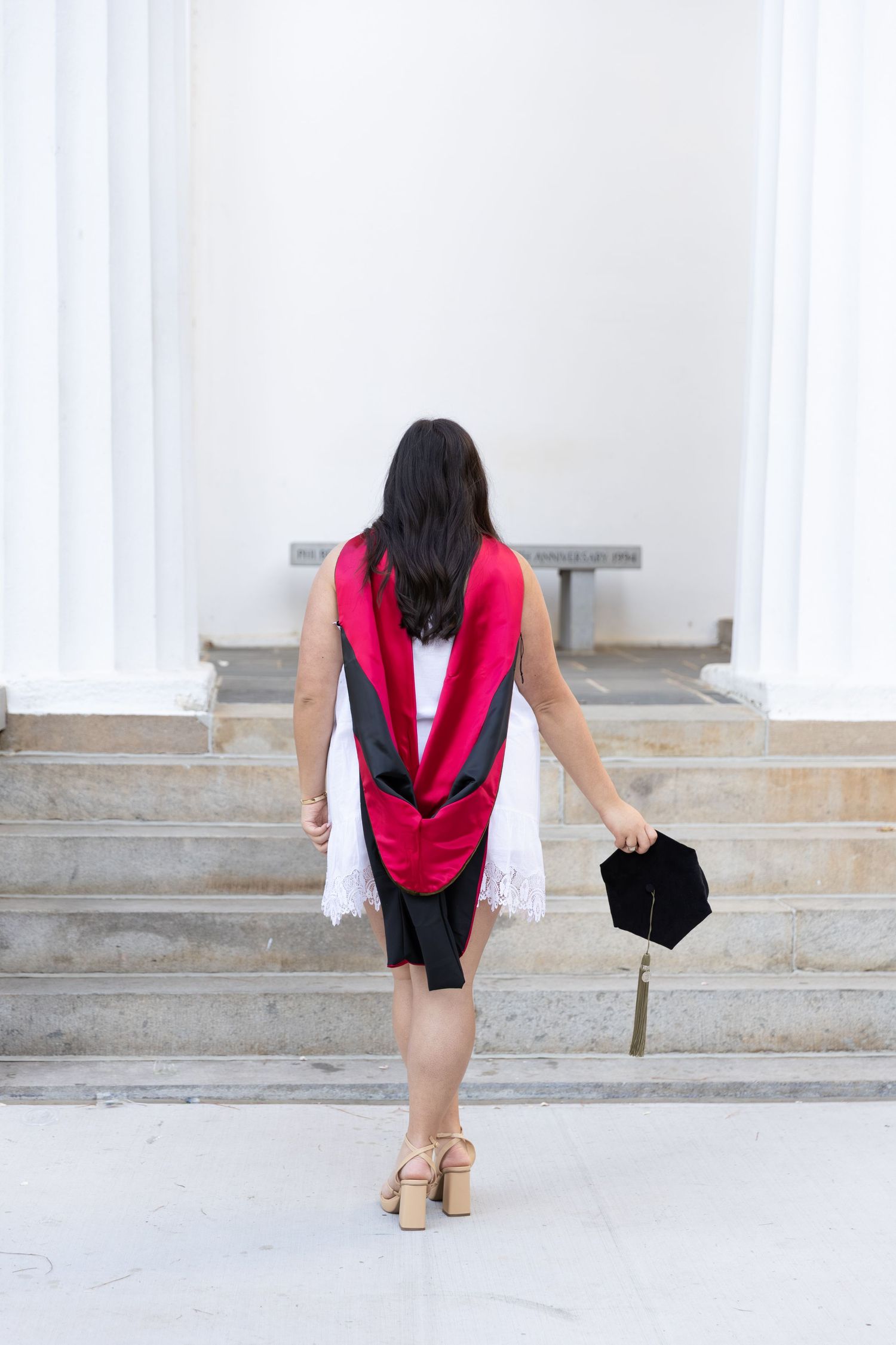 Graduate in black gown with red hood holds cap, walking toward university steps in heels.