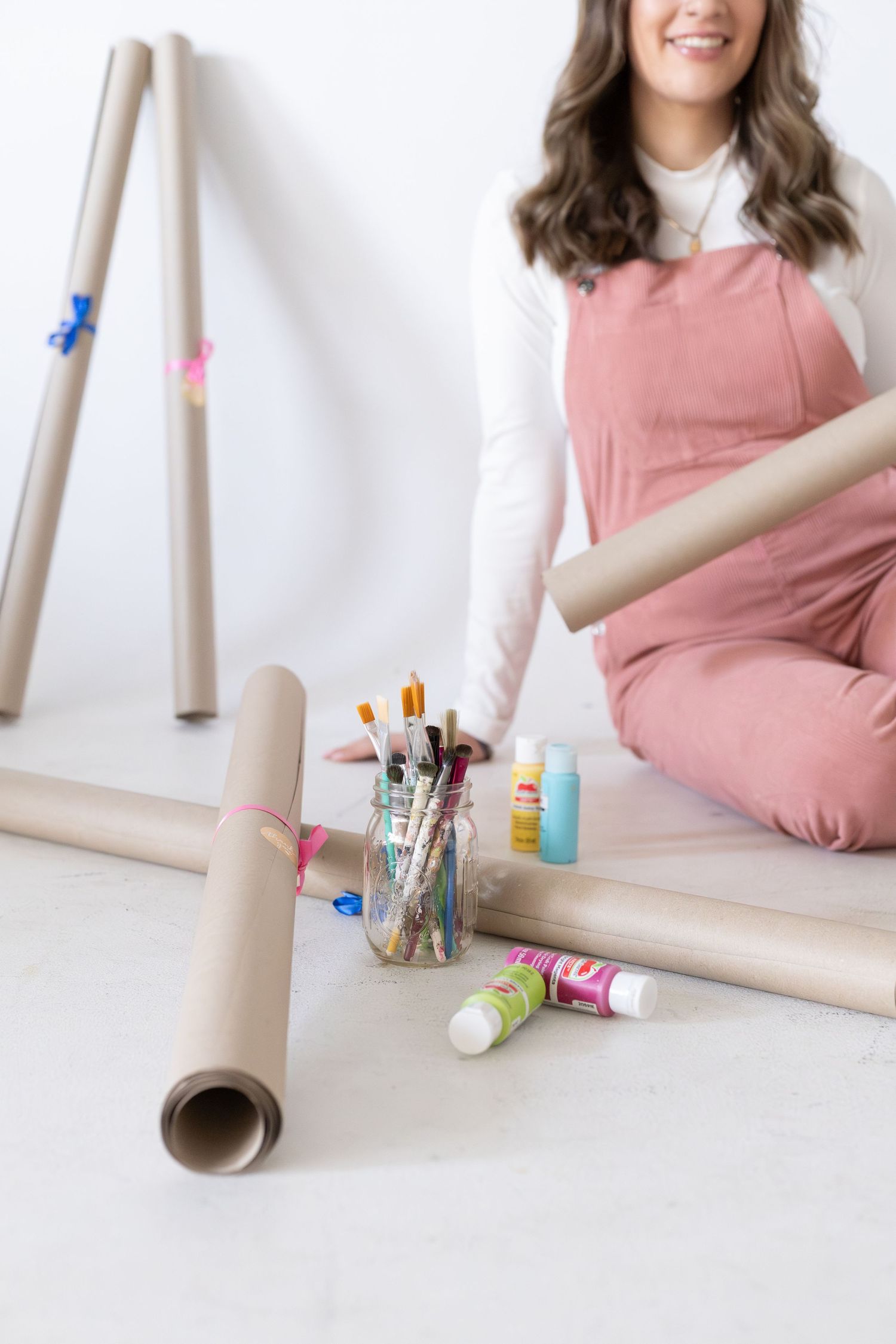 Smiling woman in pink overalls sitting on floor with paint brushes, craft tubes, and acrylic paints in bright studio.
