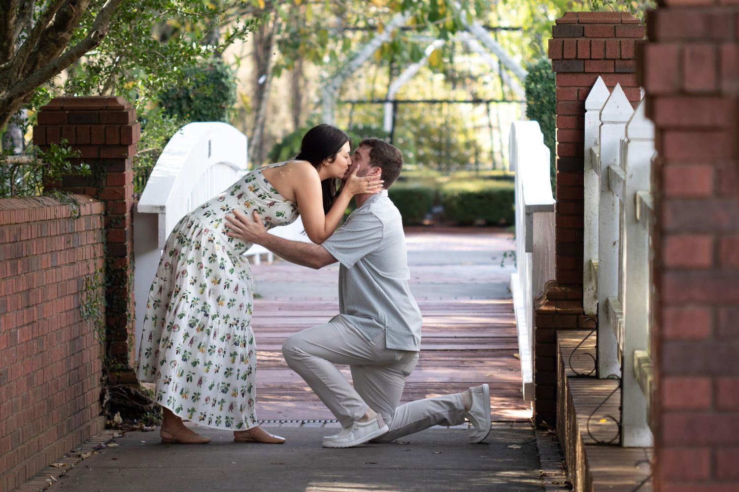 Man kneeling on one knee to kiss a woman in a floral dress near brick pillars in an outdoor garden setting.