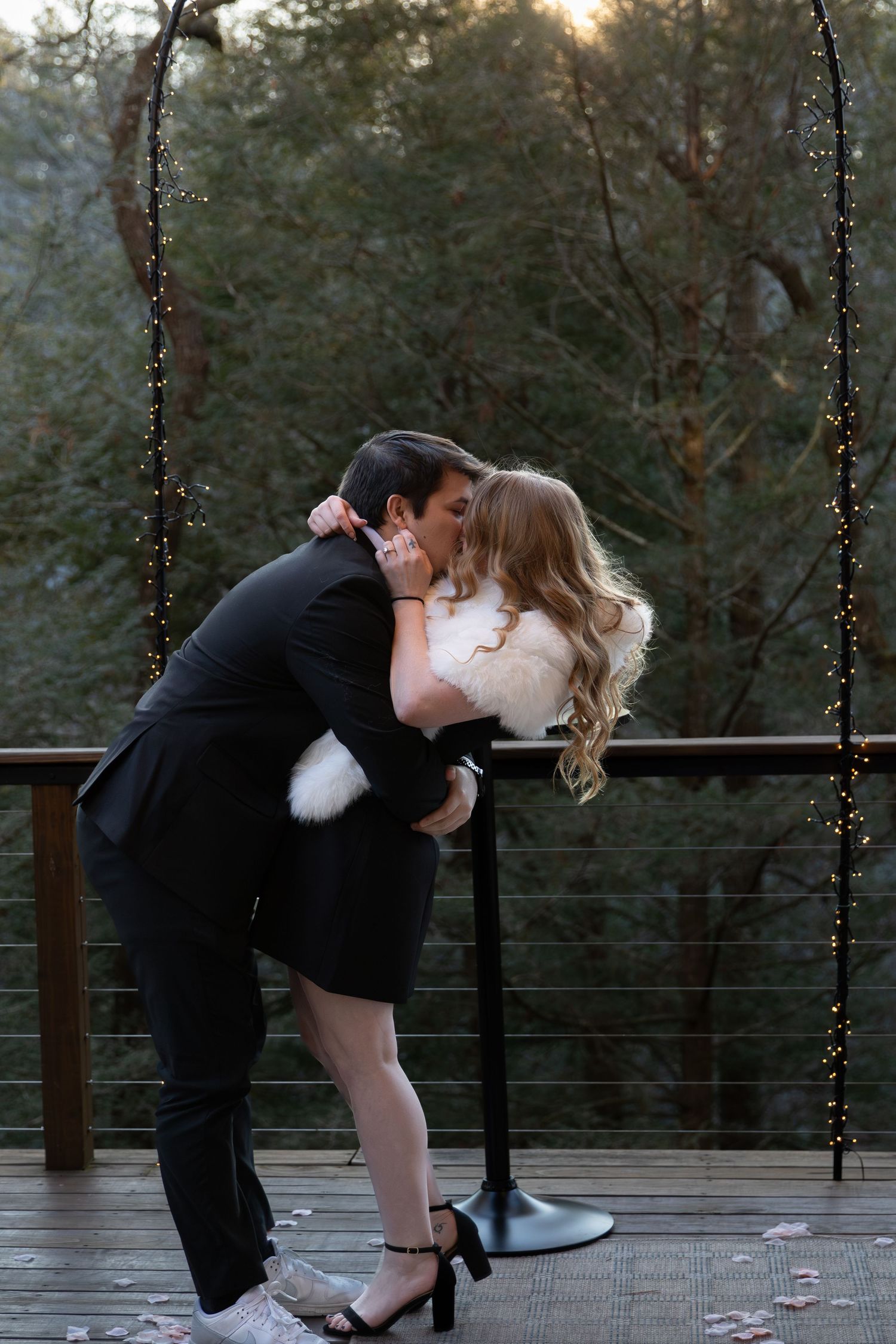 Couple sharing a romantic dip kiss on a decorated outdoor deck with fairy lights and trees in the background.