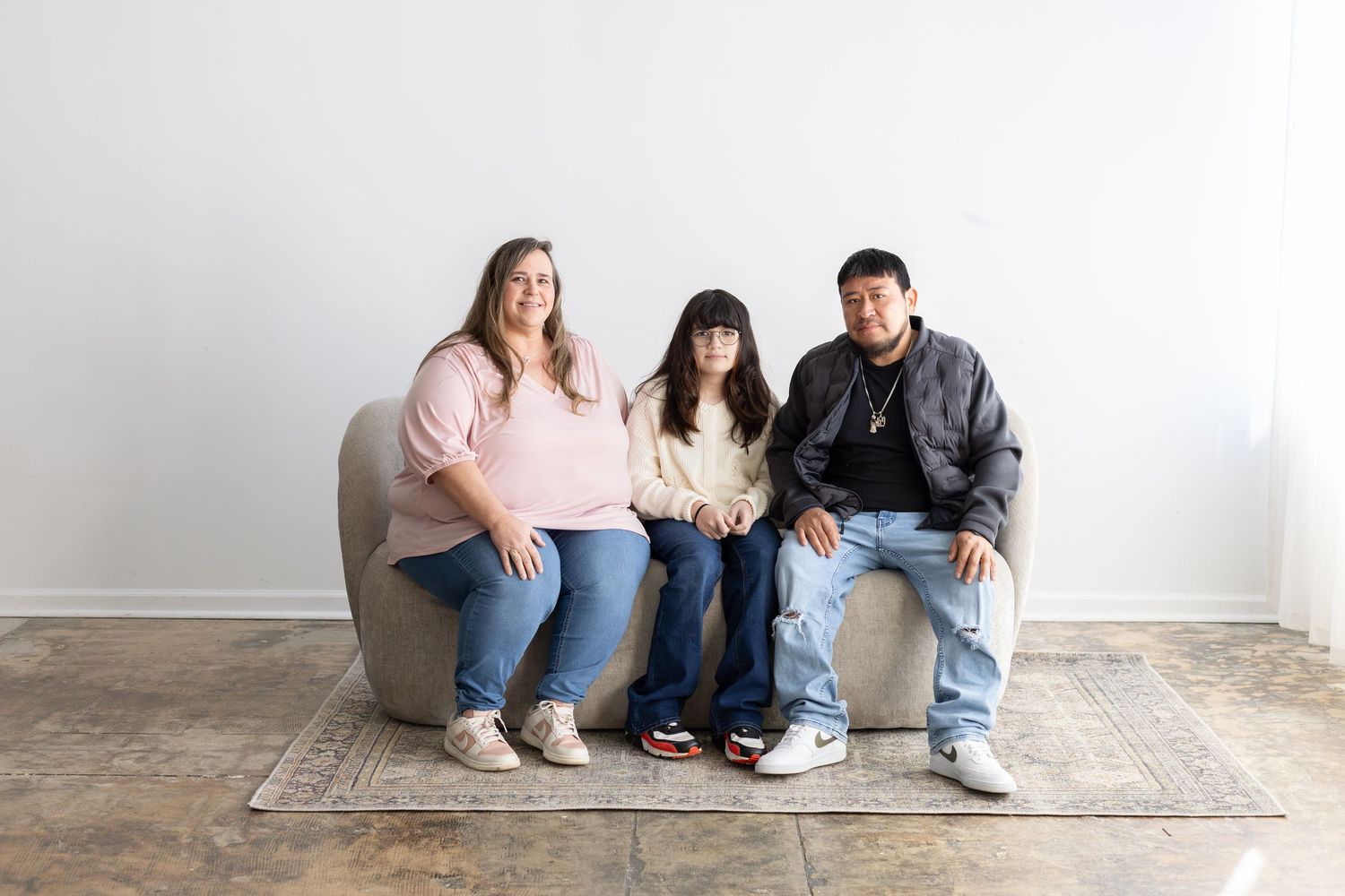 Family of three smiling together on a gray sofa against a white wall on a patterned rug.