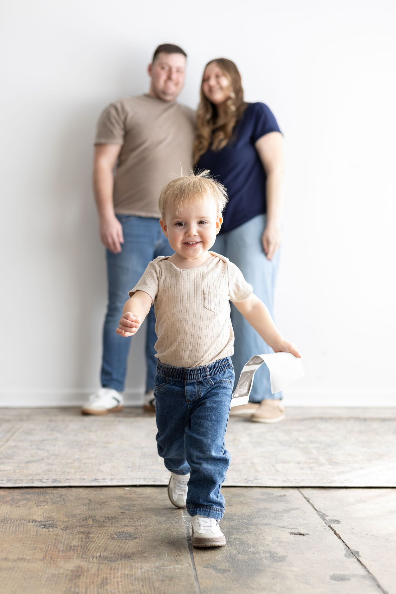 A happy toddler runs toward the camera while his parents stand smiling against a white wall behind him.