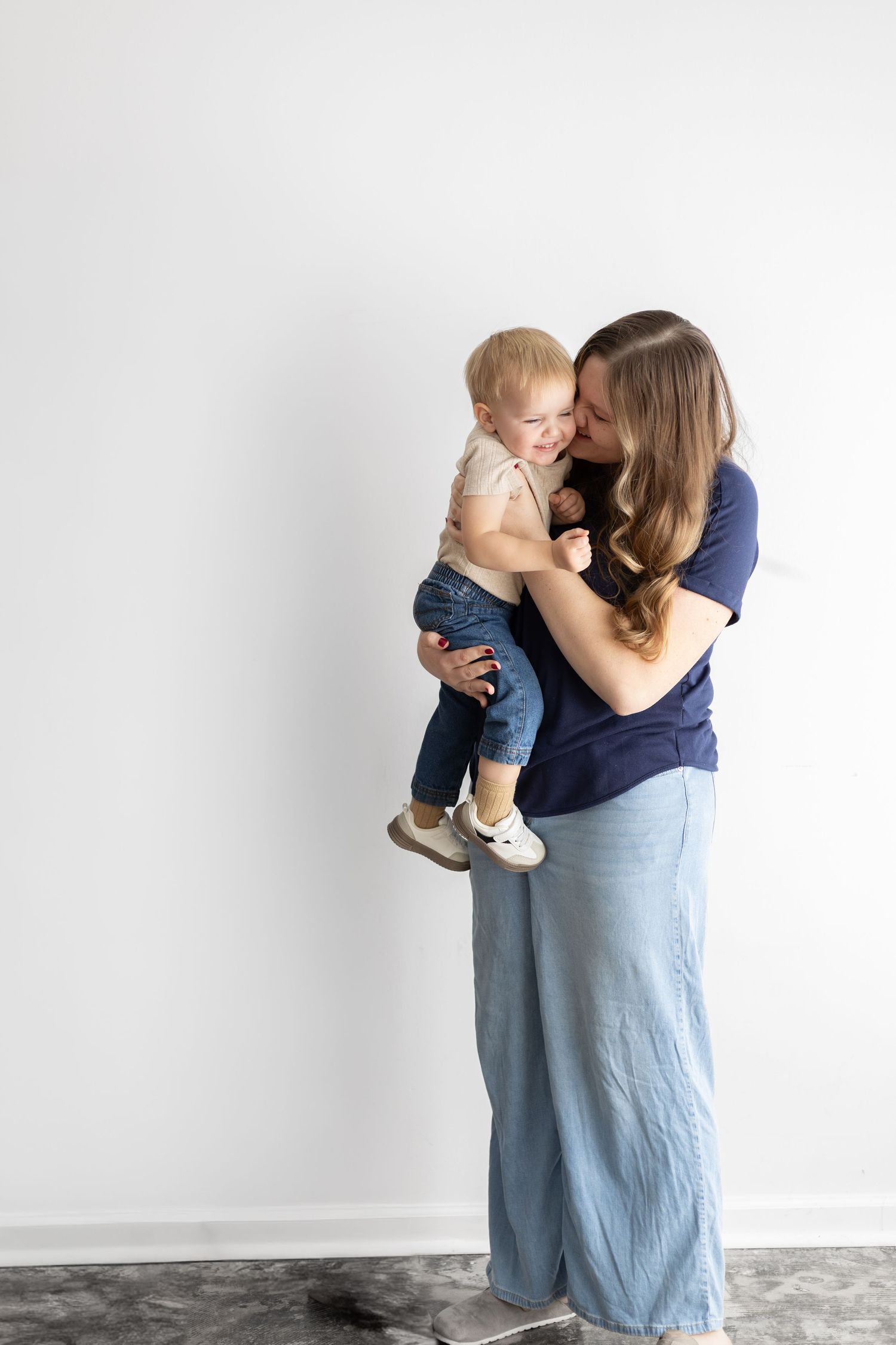 A mother lovingly holds her smiling baby while standing against a white wall, both dressed in blue.