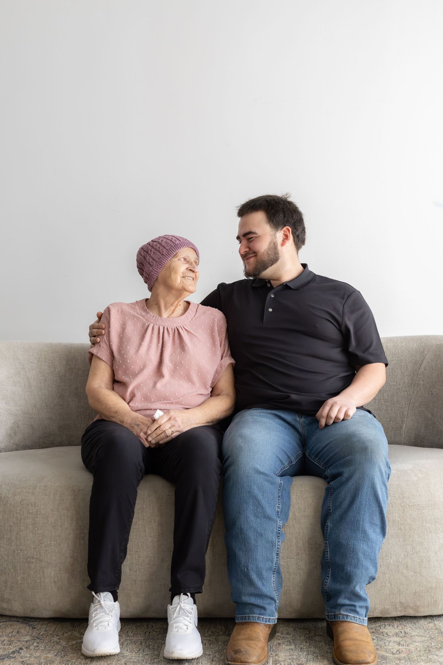 Elderly woman in pink headscarf sits with young man on couch, sharing a warm smile and embrace.