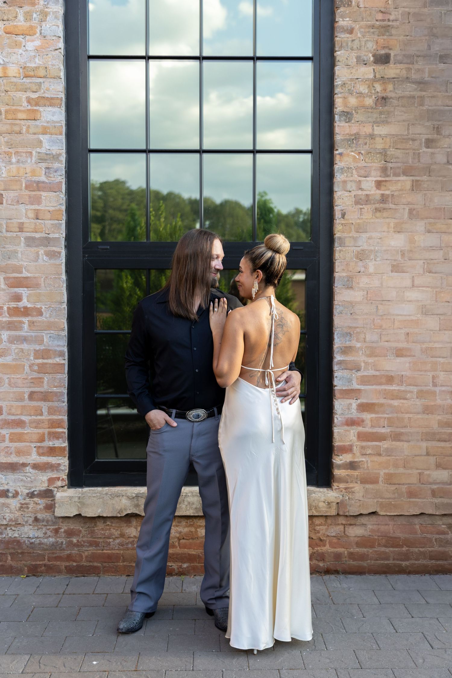 Couple in elegant attire embracing before brick wall and large window, woman in white satin gown, man in black shirt.