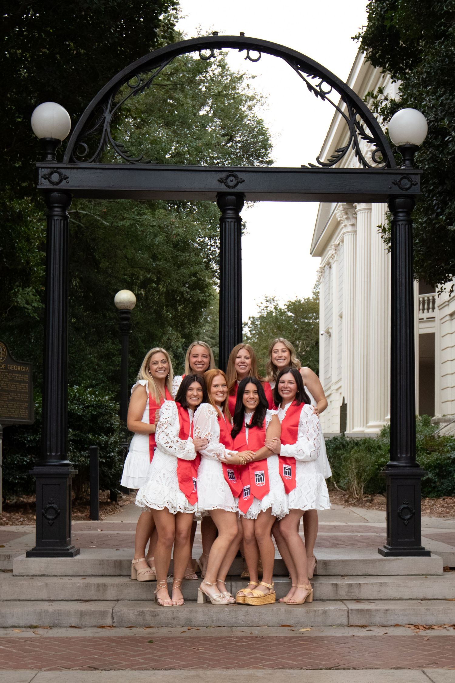 Group of women in white dresses with red sashes posing under the University of Georgia Arch landmark.