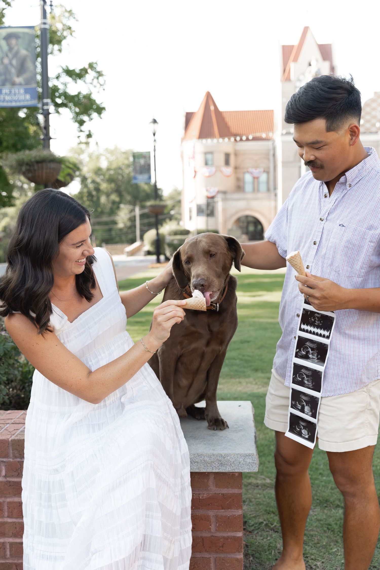 Couple in white outfits sharing ice cream with chocolate Labrador outdoors, man holding ultrasound photos.