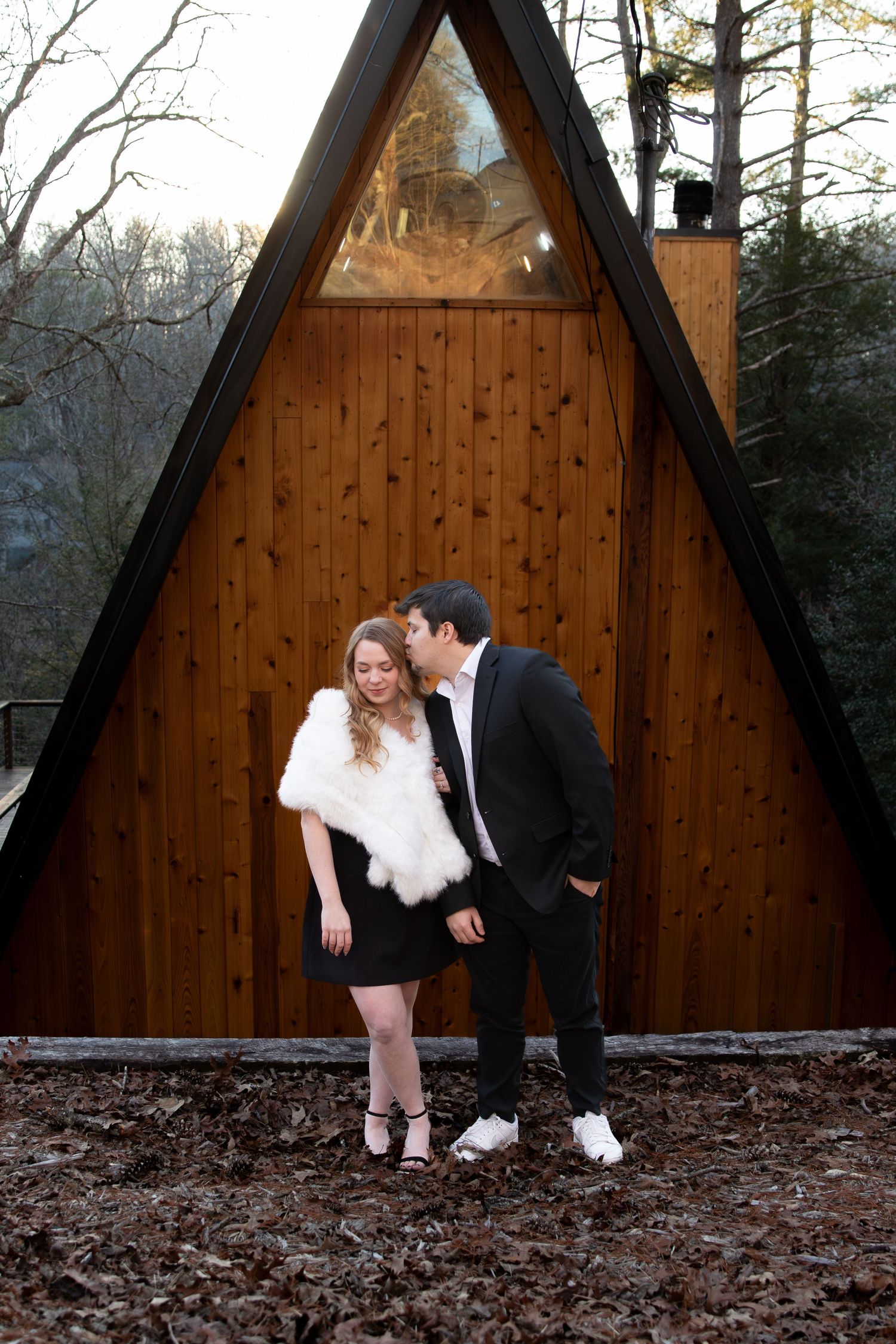 Couple posing in front of a wooden A-frame cabin, woman in white fur shawl and black dress, man in dark suit kissing her cheek.