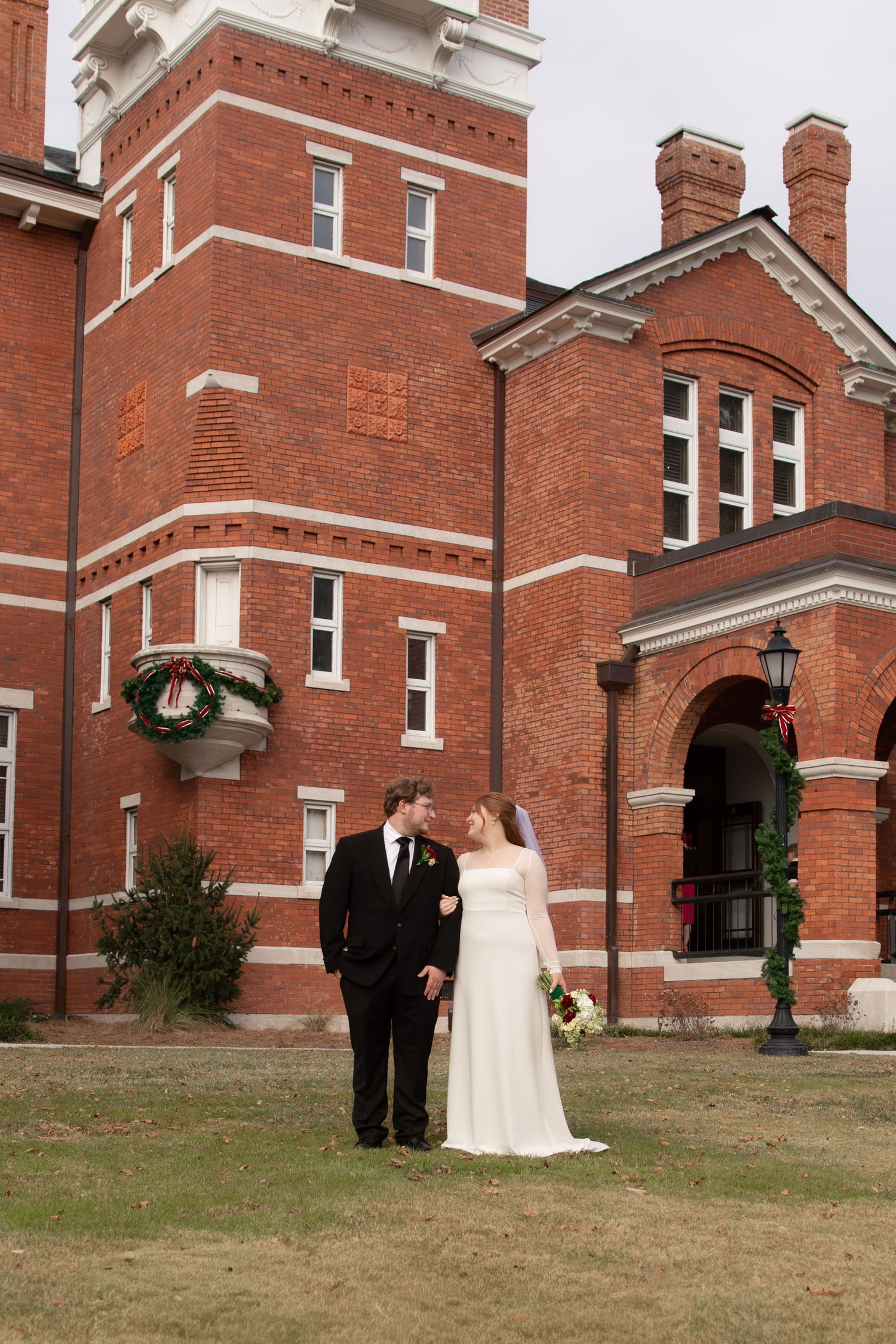 Bride and groom pose together on lawn before historic red brick building decorated with Christmas wreaths.