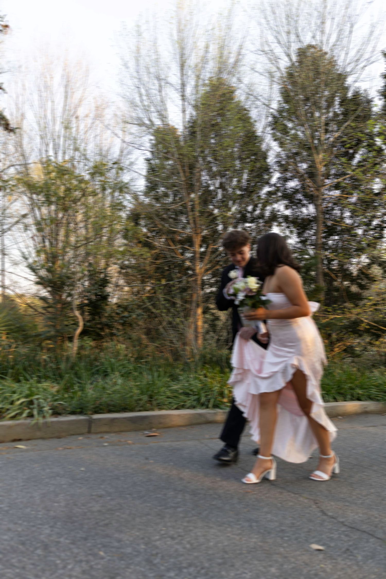 A couple in formal attire walks together outdoors, woman in white gown holding bouquet, man in black suit.