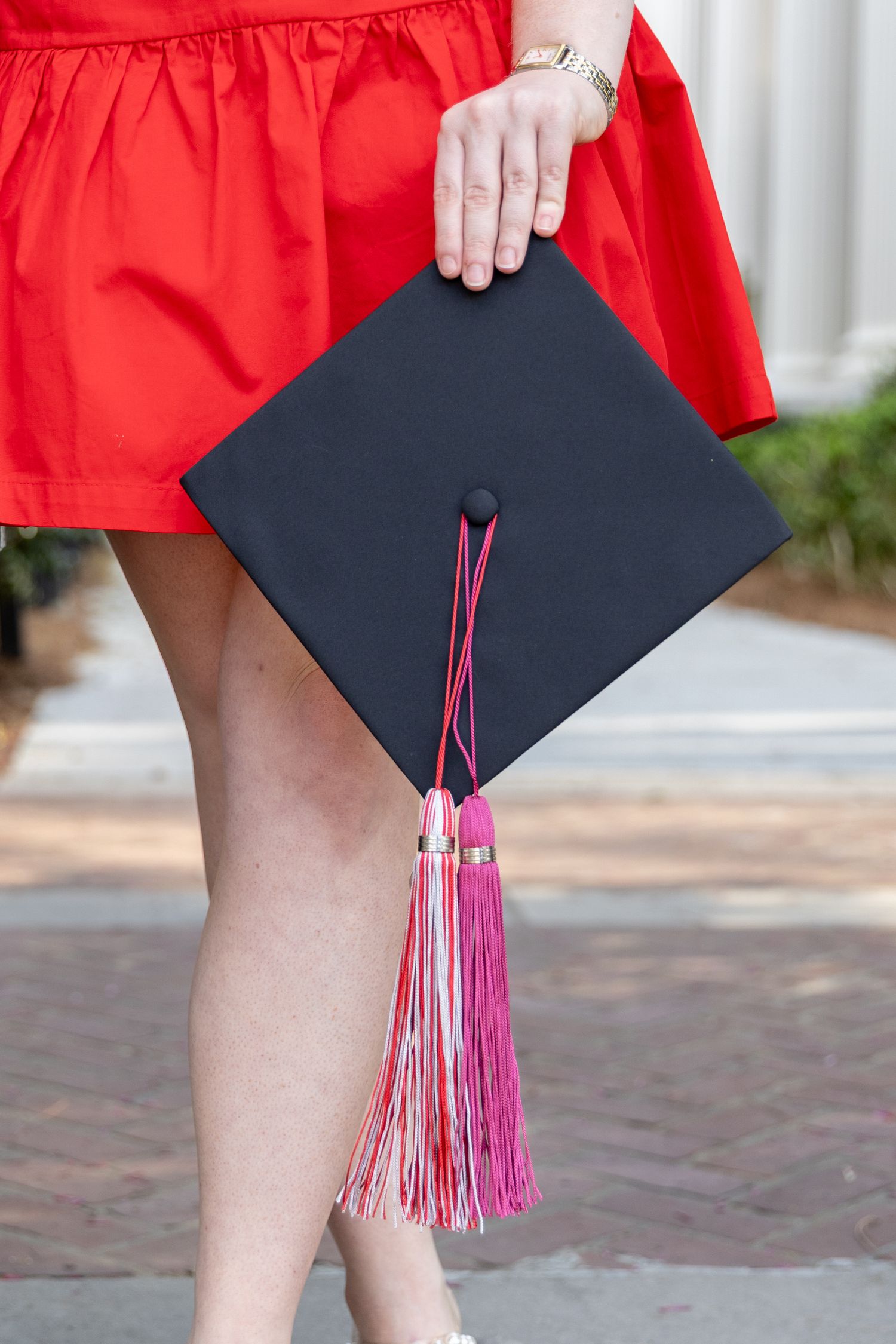 Woman in red dress holding black graduation cap with pink tassel outdoors.