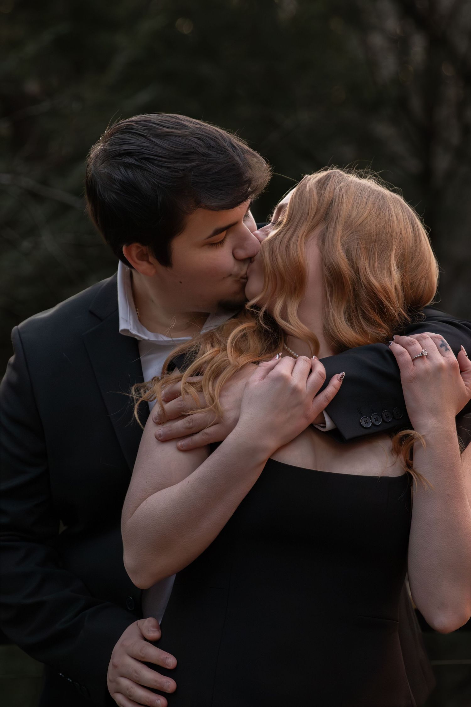 Young couple kissing and embracing outdoors, dressed in elegant black formal attire.