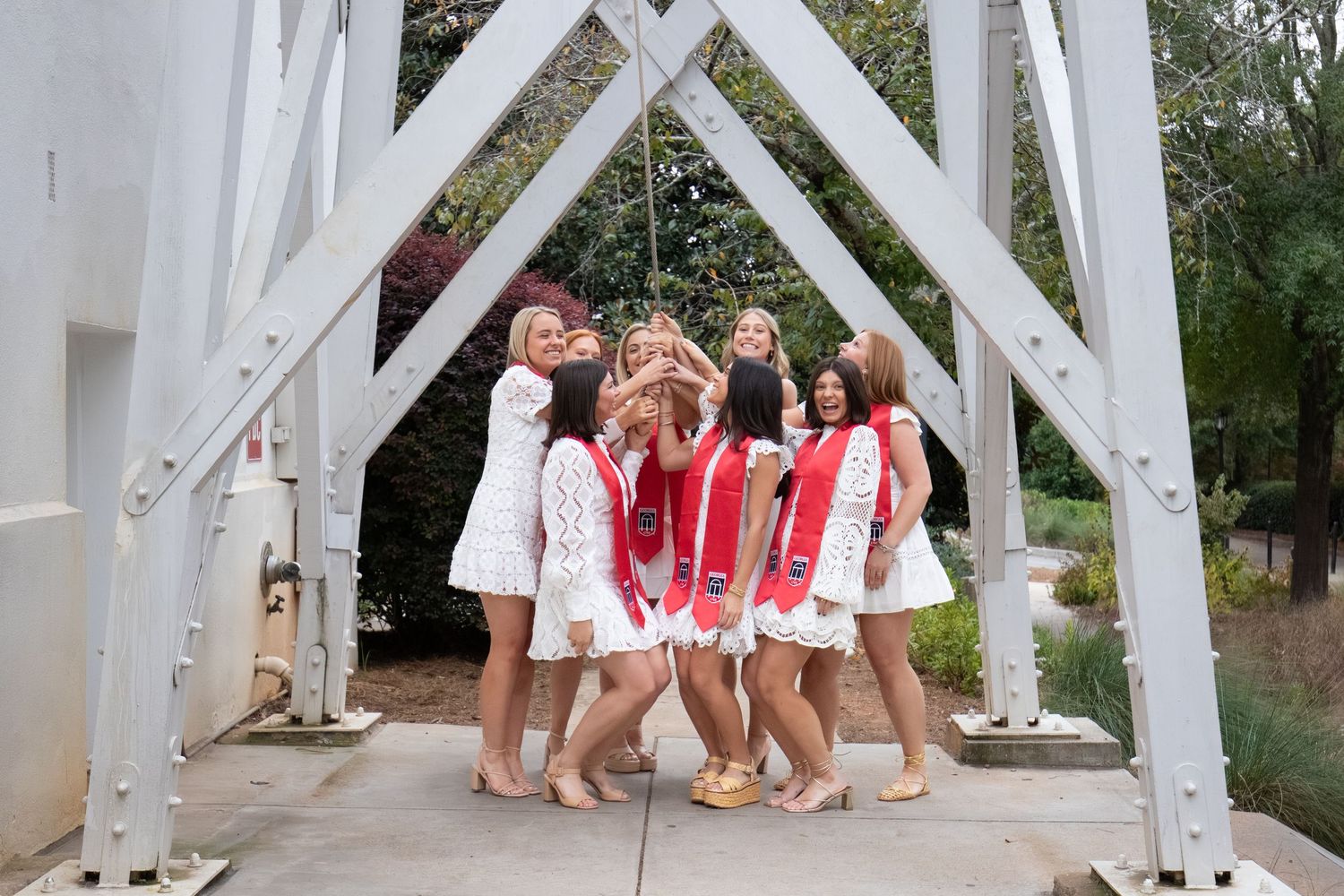Group of young women in white dresses celebrating on a bridge, one wearing red graduation sashes.