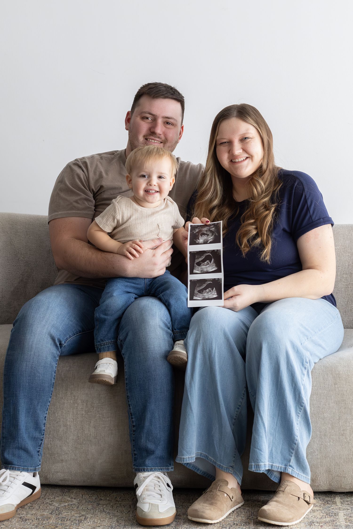 Happy family of three sitting on a couch holding ultrasound photos, announcing a second pregnancy.