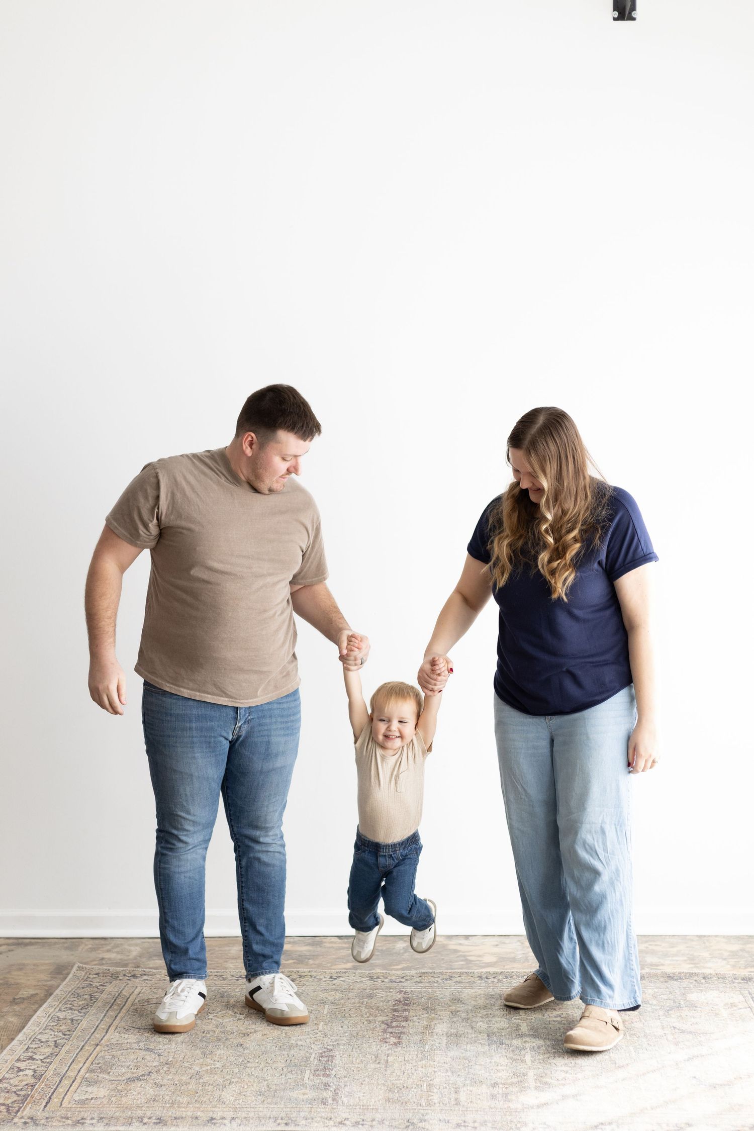 Parents joyfully hold their laughing toddler's hands as he takes steps between them in a bright white room.