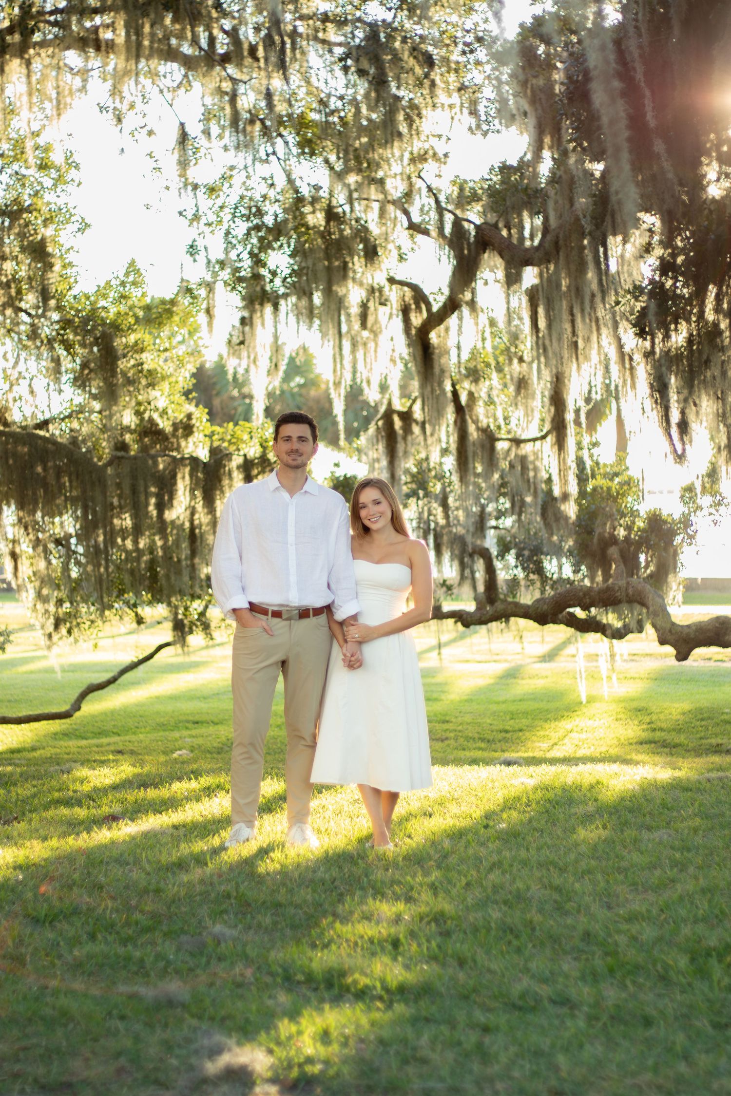 Couple in white and khaki attire holding hands on green lawn beneath Spanish moss-draped oak trees in golden sunlight.