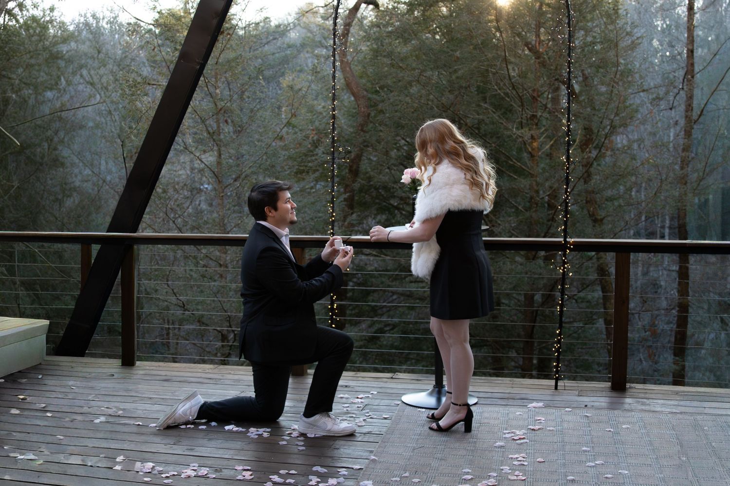 Man kneeling on outdoor deck proposing to woman with forest backdrop and string lights.