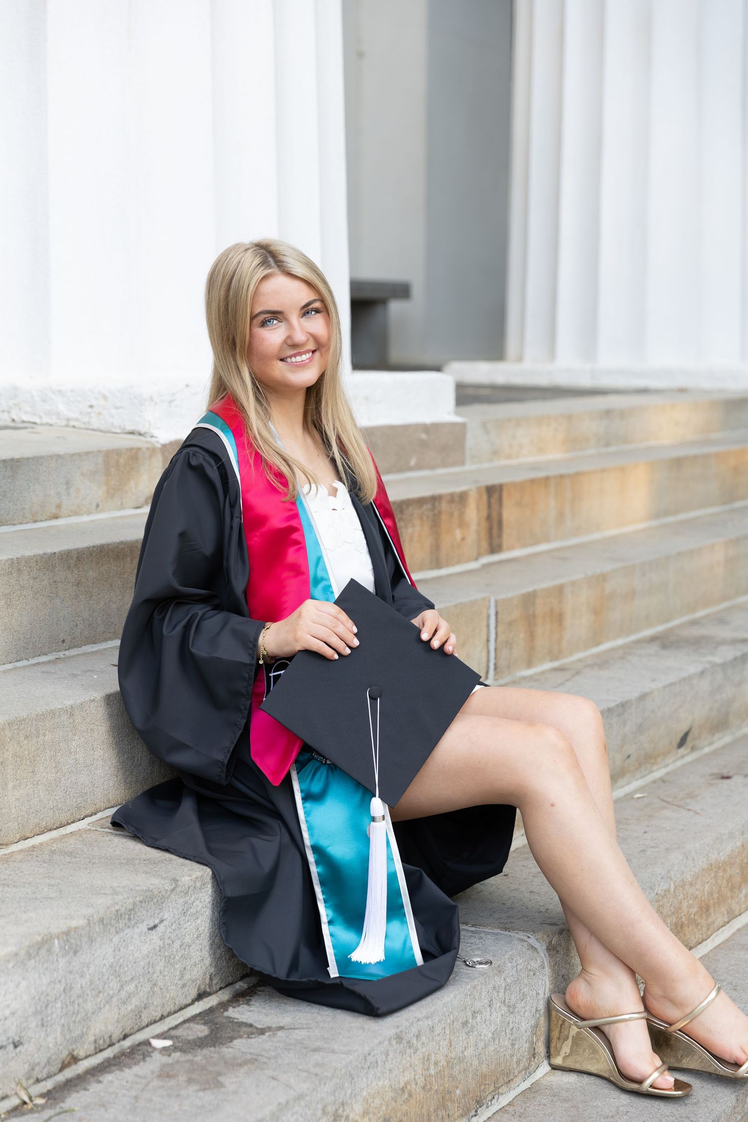 Smiling blonde woman in black graduation gown with colorful sashes sitting on stone steps holding mortarboard cap.