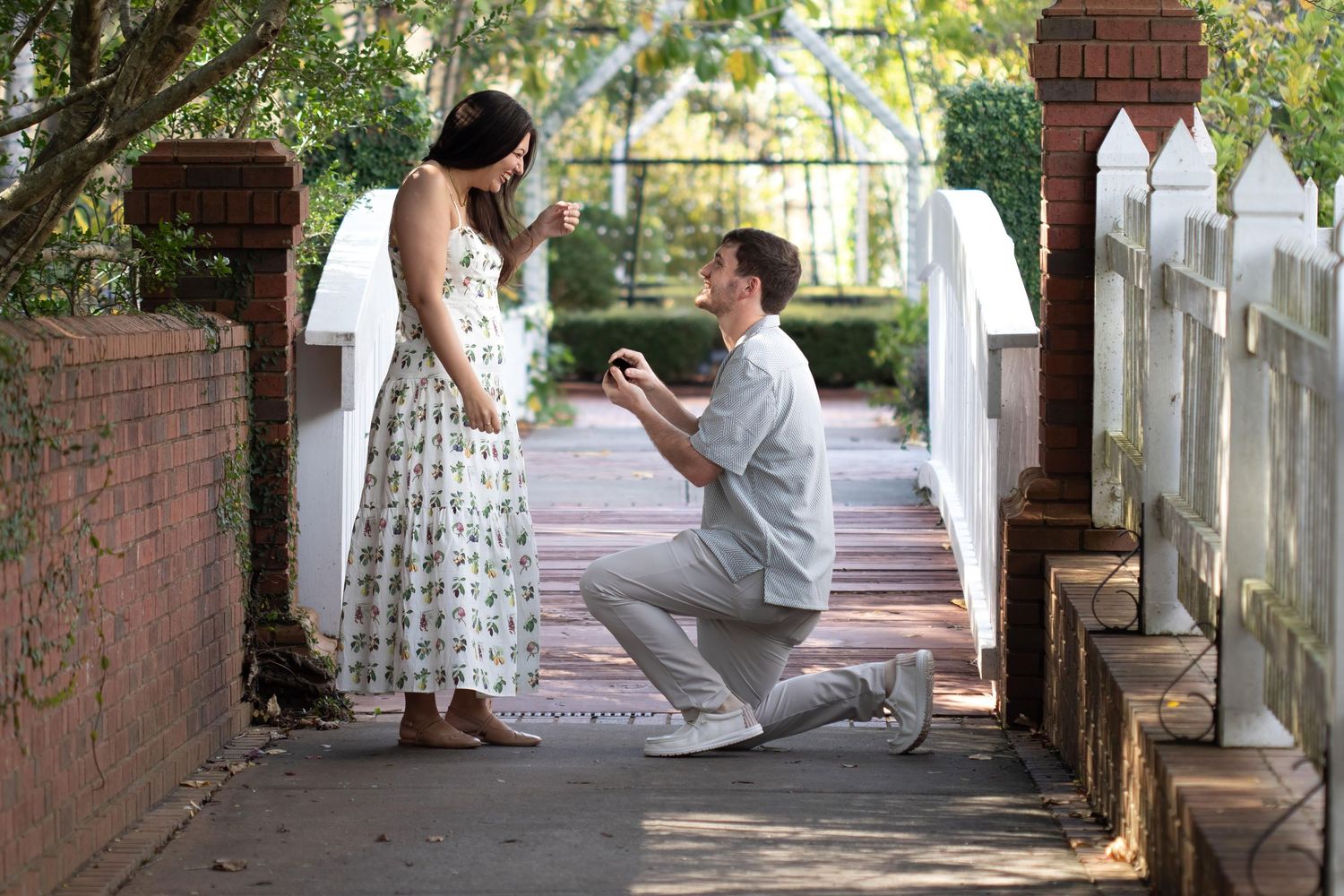 Man kneeling on one knee proposing to woman in floral dress on a charming brick pathway surrounded by lush greenery.