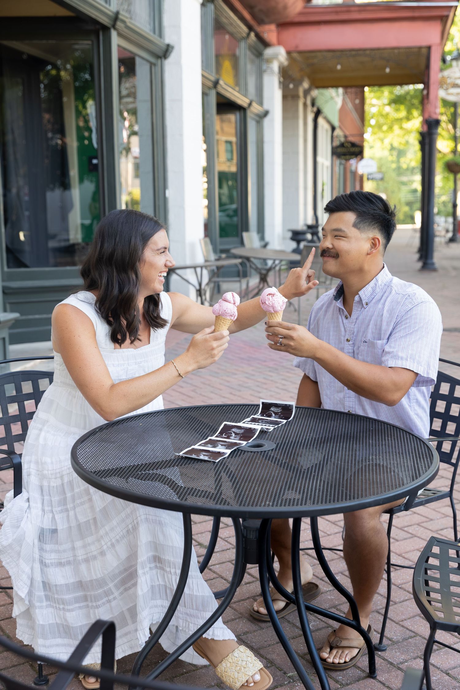 Happy couple toasting ice cream cones at outdoor cafe table with ultrasound photos, celebrating pregnancy announcement.
