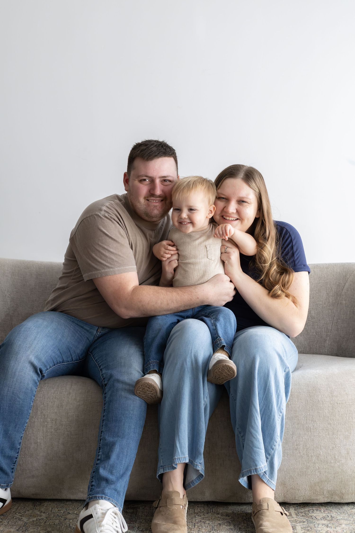 Happy young family with baby sitting together on a gray sofa, smiling at camera in a bright room.
